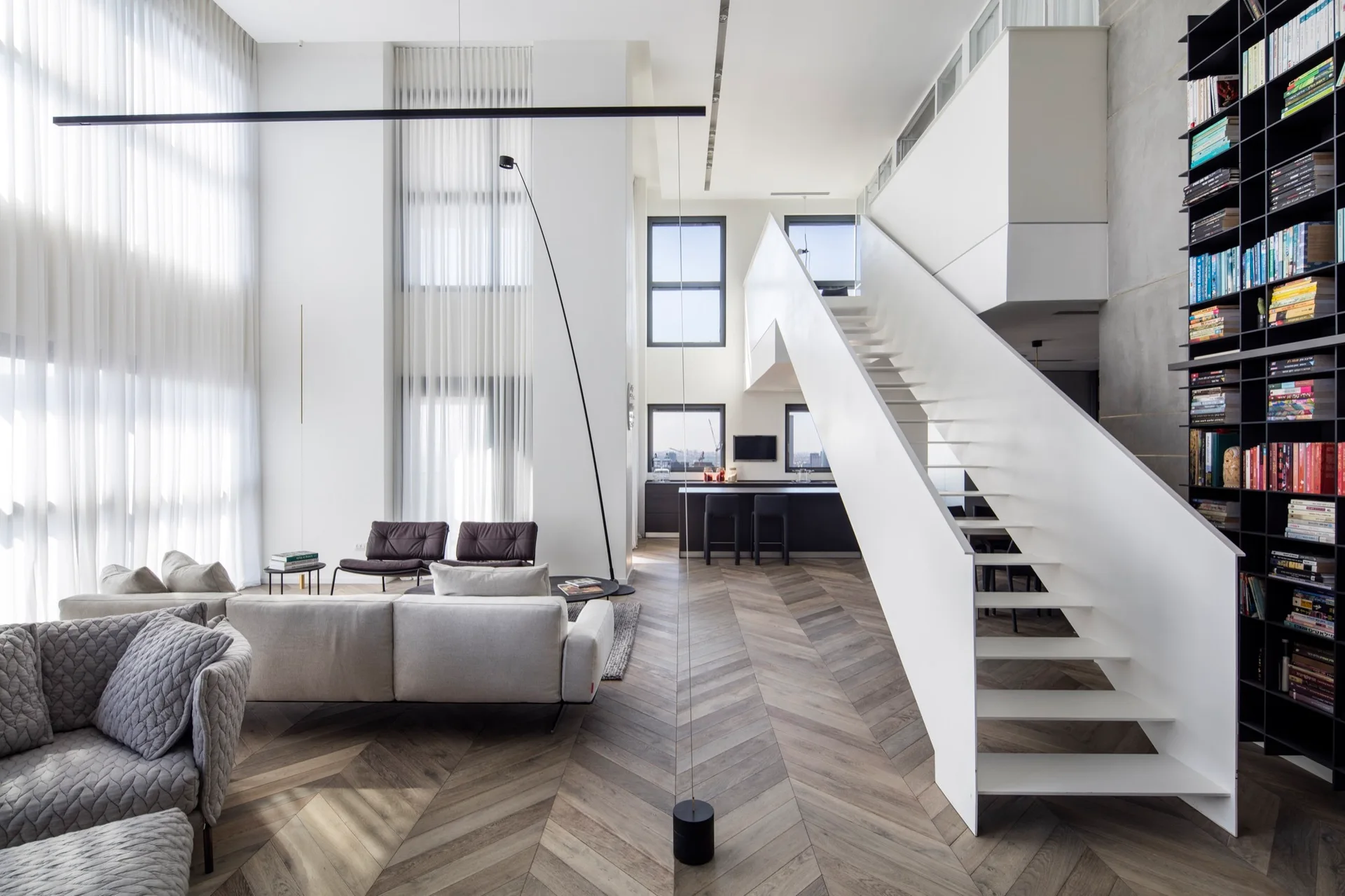 A spacious double-height living room features a white modern staircase, herringbone wood floors, a cream sectional sofa, and a tall black bookshelf.