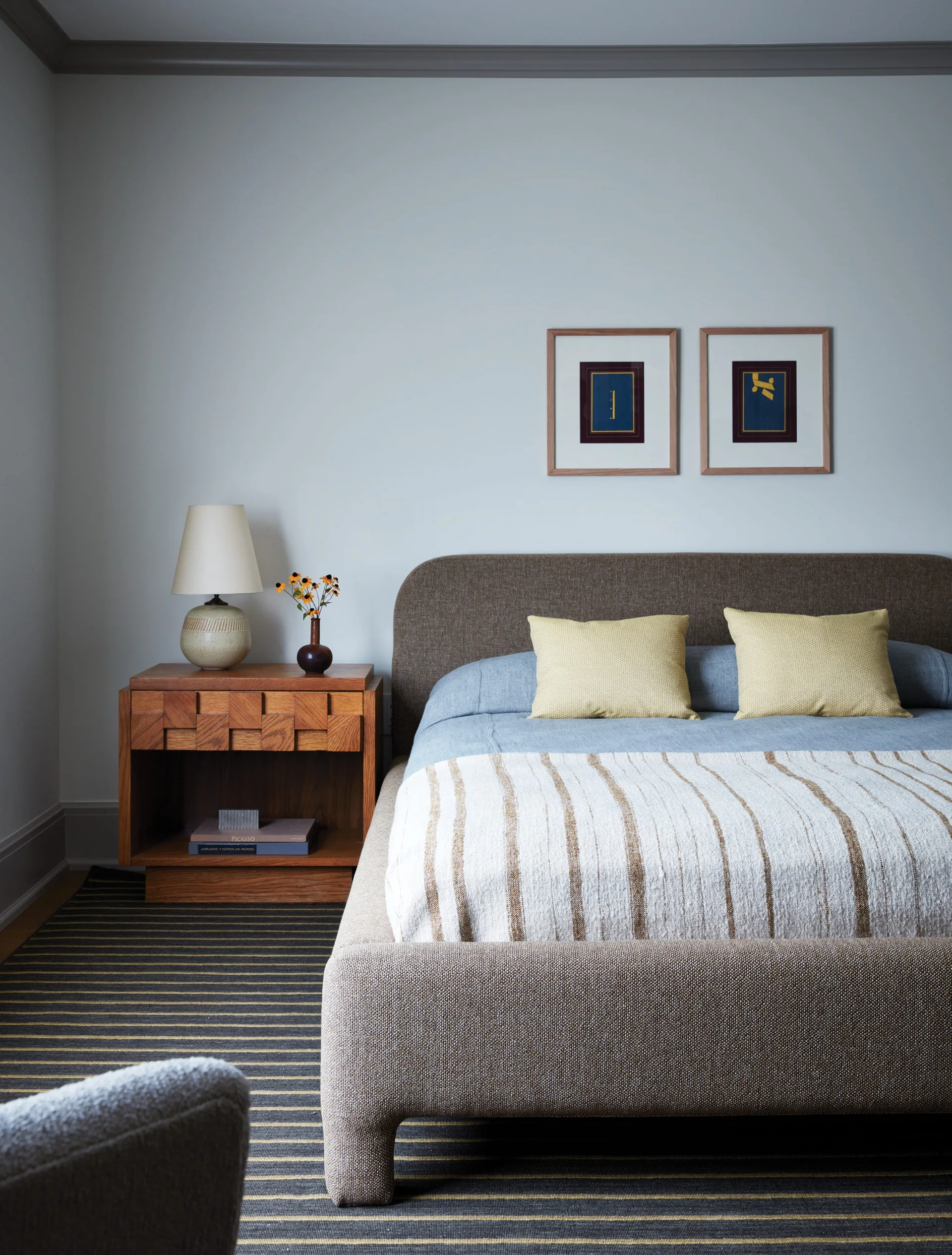 A bedroom features a brown upholstered bed with blue bedding, a geometric wood nightstand with a lamp, and a striped rug.