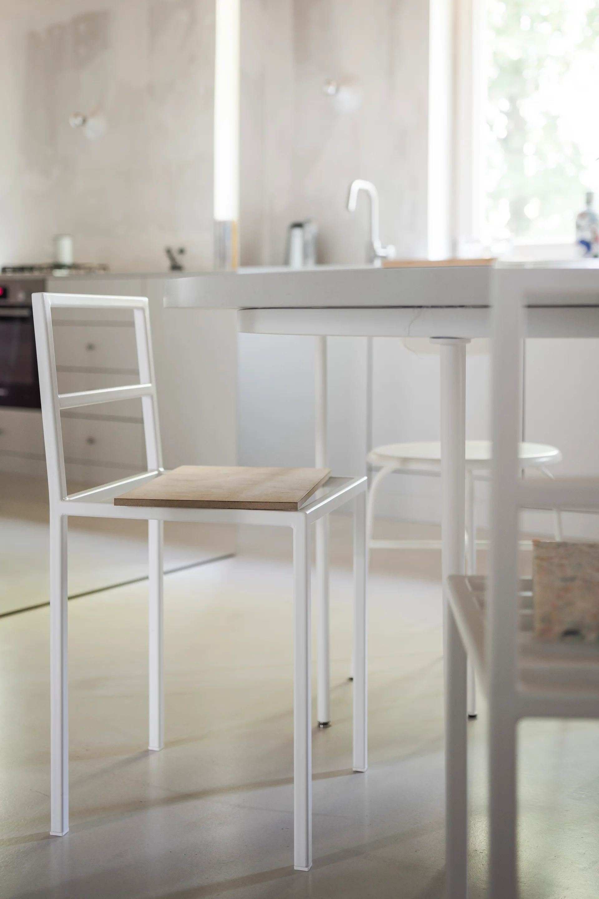 A minimalist white metal dining chair with a natural wood seat sits next to a white table in a bright, modern kitchen.