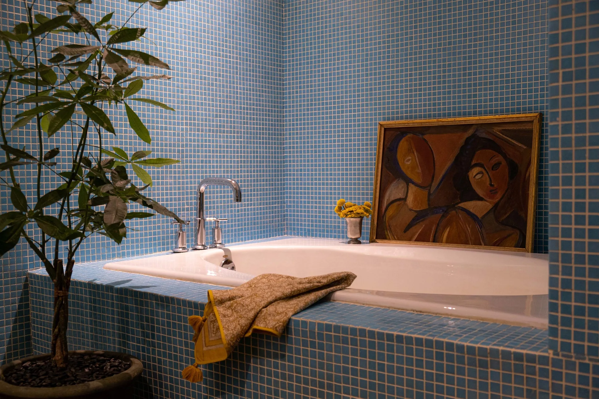 A white drop-in bathtub is set into a platform covered in blue mosaic tile next to a framed painting and a potted plant.