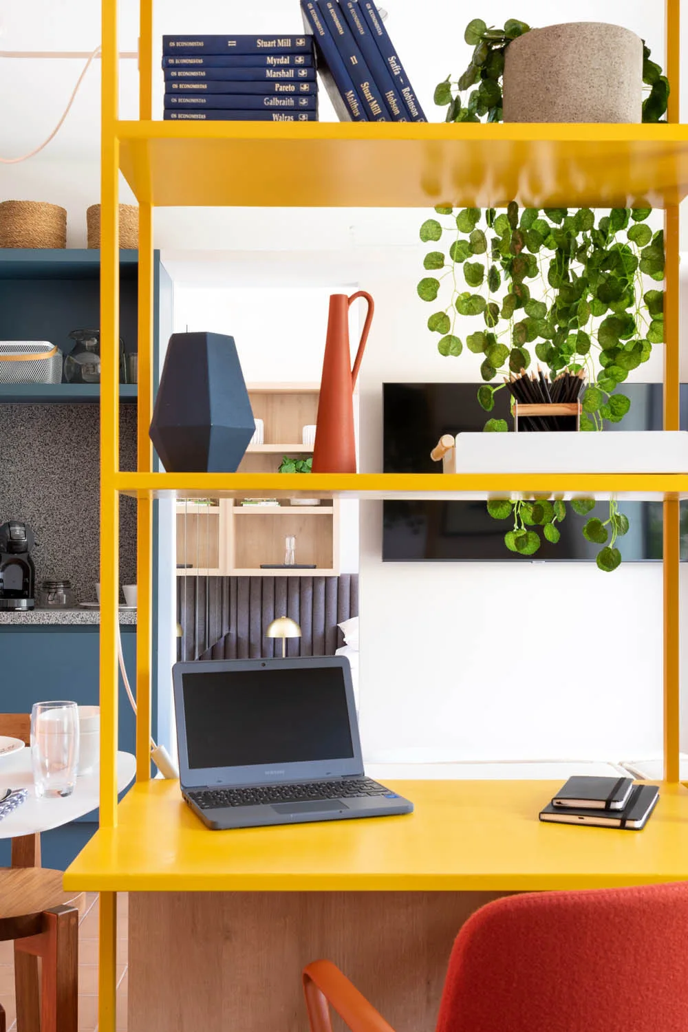 A bright yellow open shelving unit with a laptop on the desk shelf stands next to an orange chair.