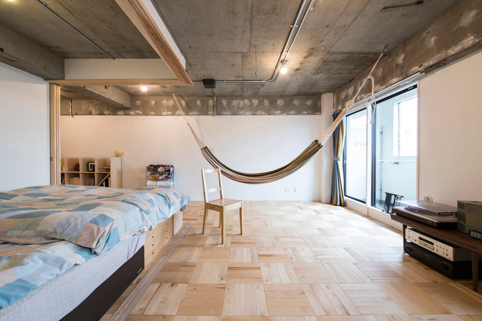 A minimalist bedroom featuring a low platform bed, a hanging fabric hammock, light wood parquet flooring, and an exposed concrete ceiling.