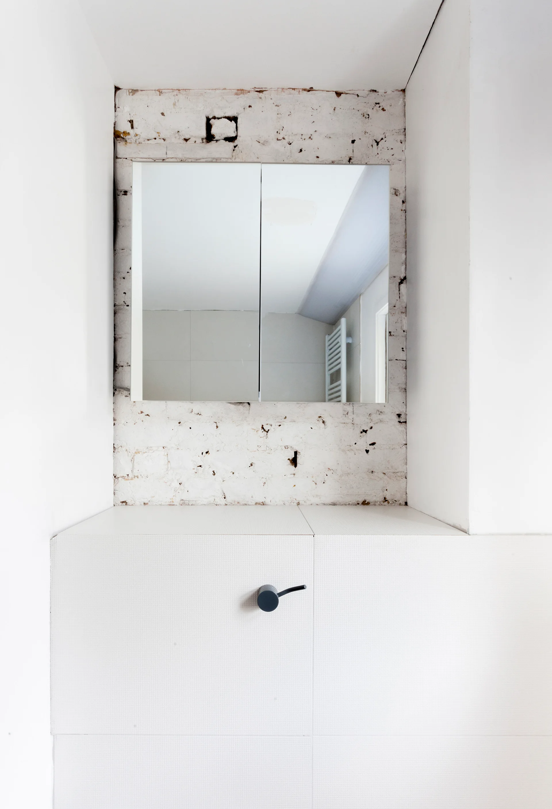 A minimalist white bathroom vanity and frameless rectangular mirror set inside a recessed alcove with a white-painted exposed brick wall.