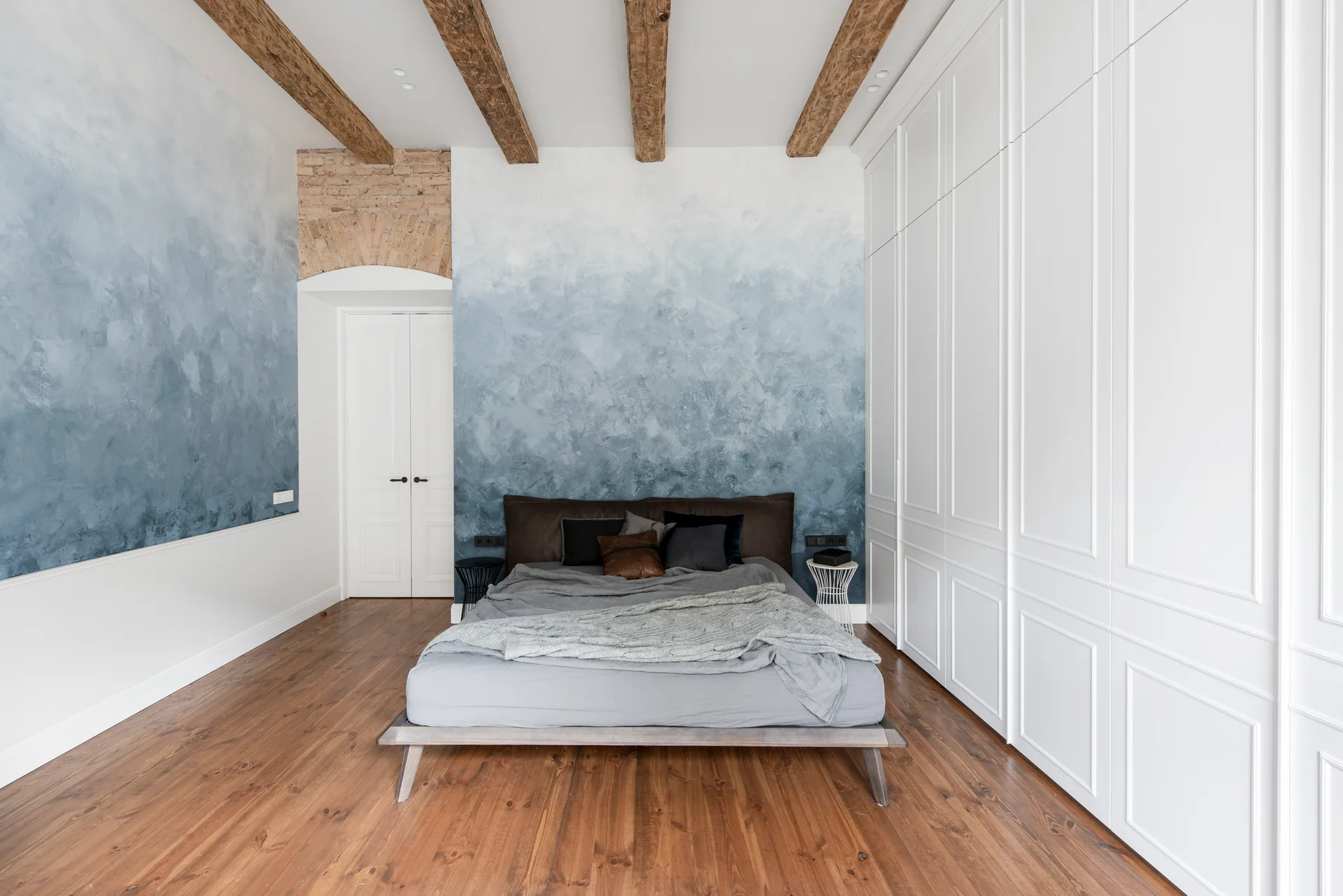 A modern bedroom featuring a low-profile bed against a textured blue ombre wall with rustic ceiling beams and built-in white wardrobes.