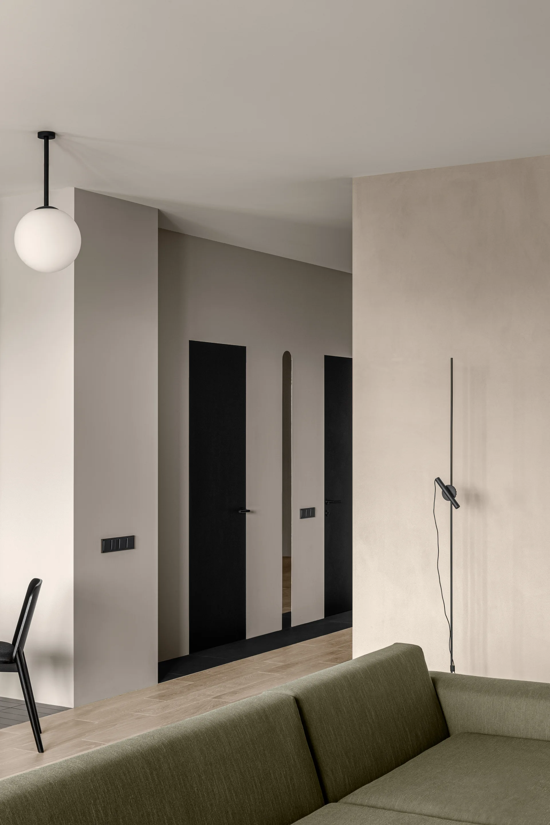 An olive green fabric sofa rests in a minimalist living room featuring smooth beige plaster walls, a globe pendant light, and striking matte black doors.
