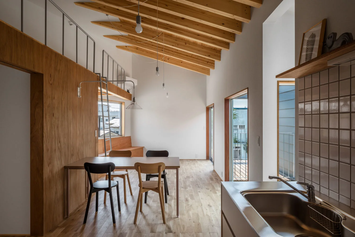 An open plan kitchen and dining area with a vaulted wood beam ceiling, wood-paneled wall, dining table with mismatched chairs, and a stainless steel sink.