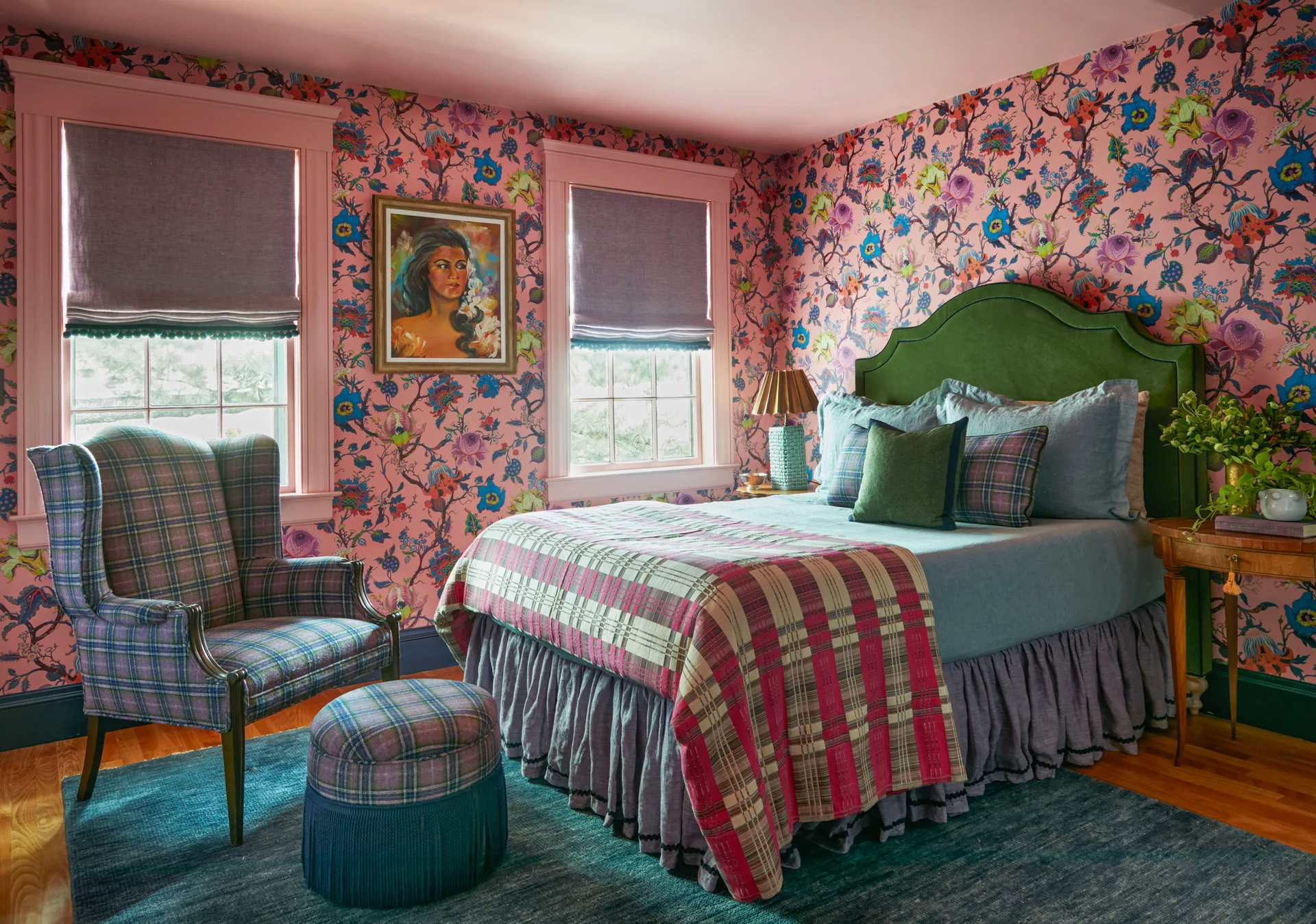 A bedroom features bright pink floral wallpaper, a green upholstered headboard, a plaid wingback chair, and matching plaid ottoman on a blue rug.