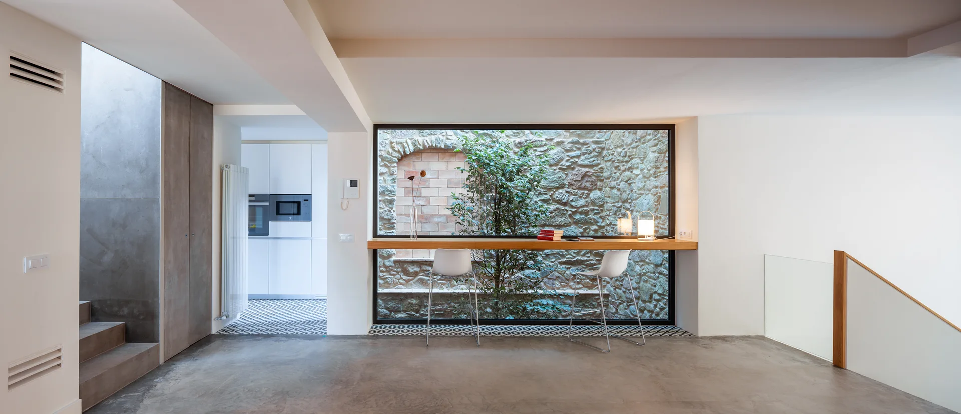 A modern living area with concrete floors features a wooden bar counter facing a large window that looks onto an outdoor stone wall.