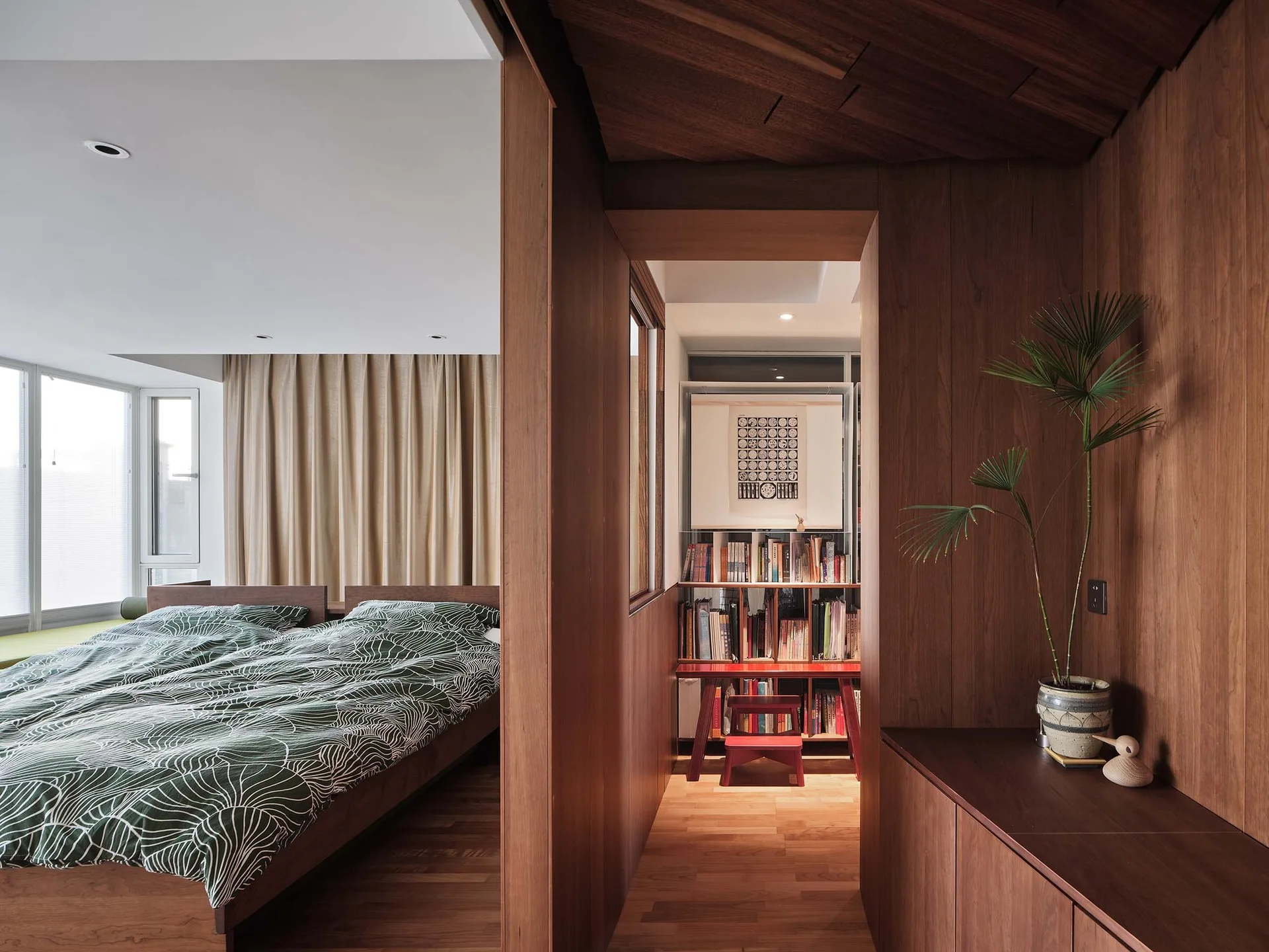 A minimalist bedroom with green botanical bedding next to a warm wood-paneled hallway containing a built-in bookshelf and a red bench.
