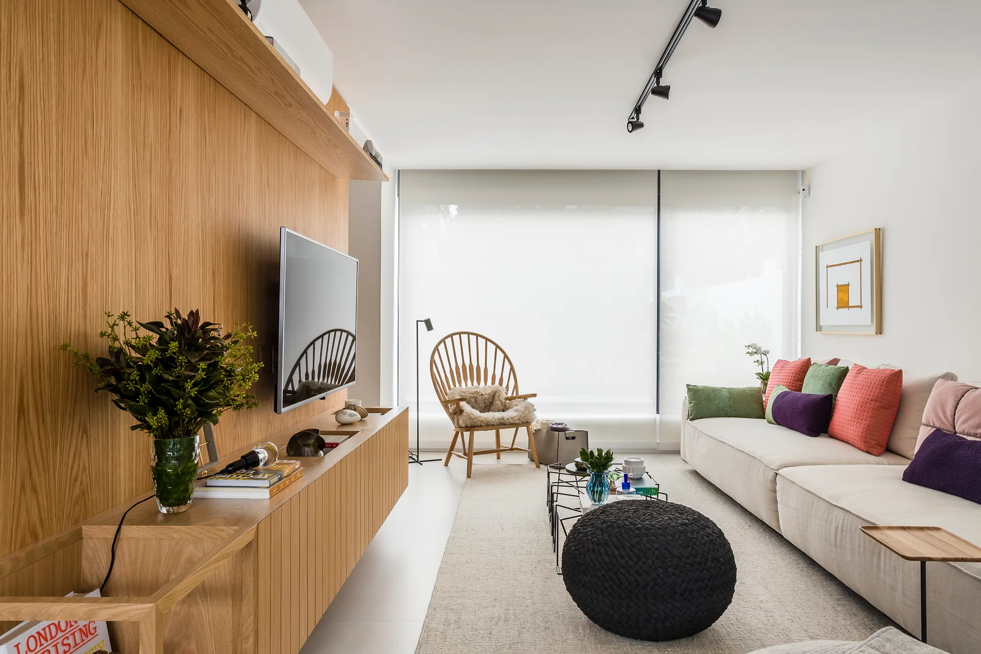 A modern living room featuring a light oak wood paneled wall with a mounted television, a cream sofa with colorful pillows, and a wood spindle chair.