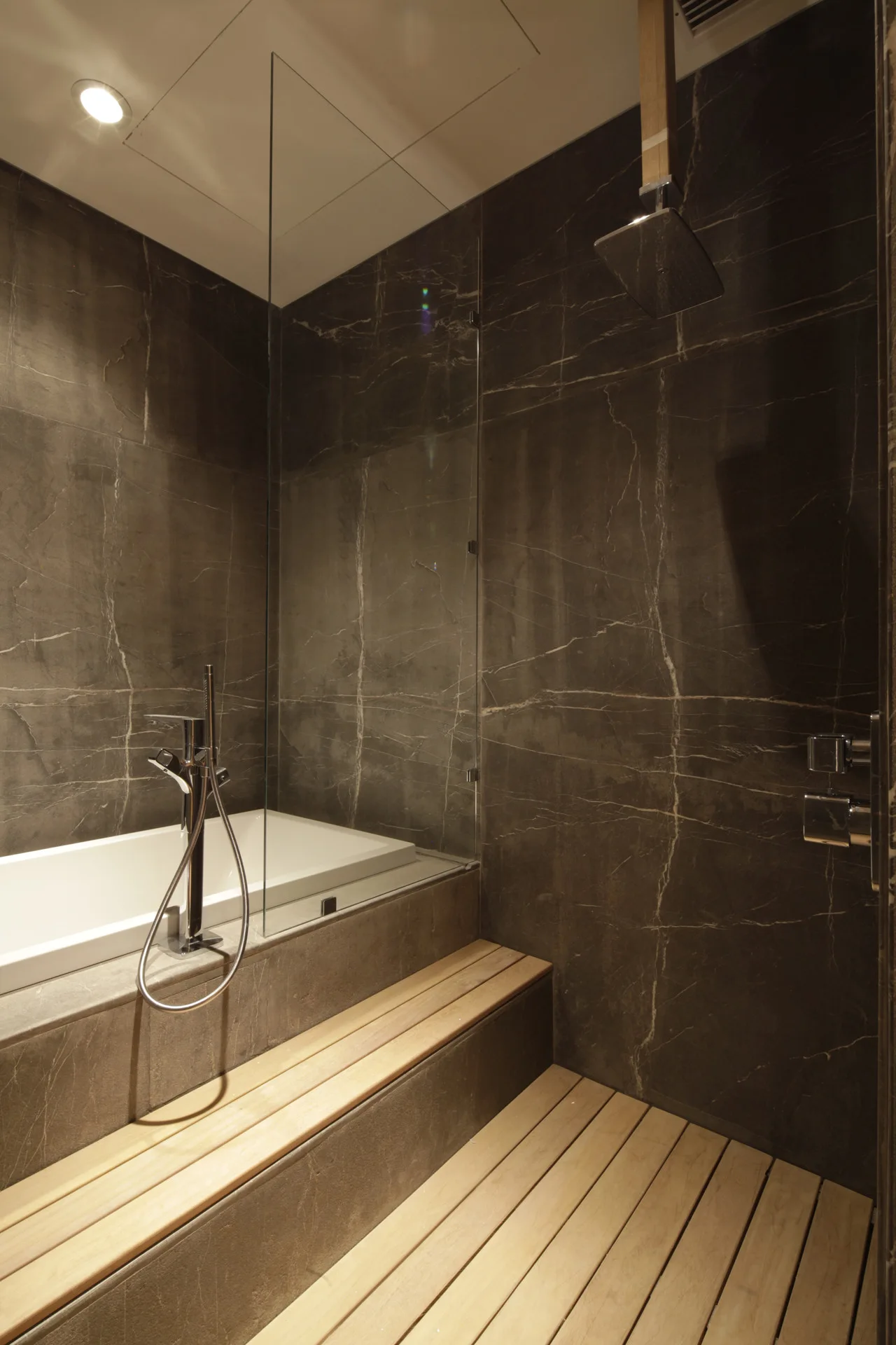 A modern bathroom featuring dark charcoal marble-look wall tiles, a white bathtub with a glass partition, and a light wood slatted bench and floor.