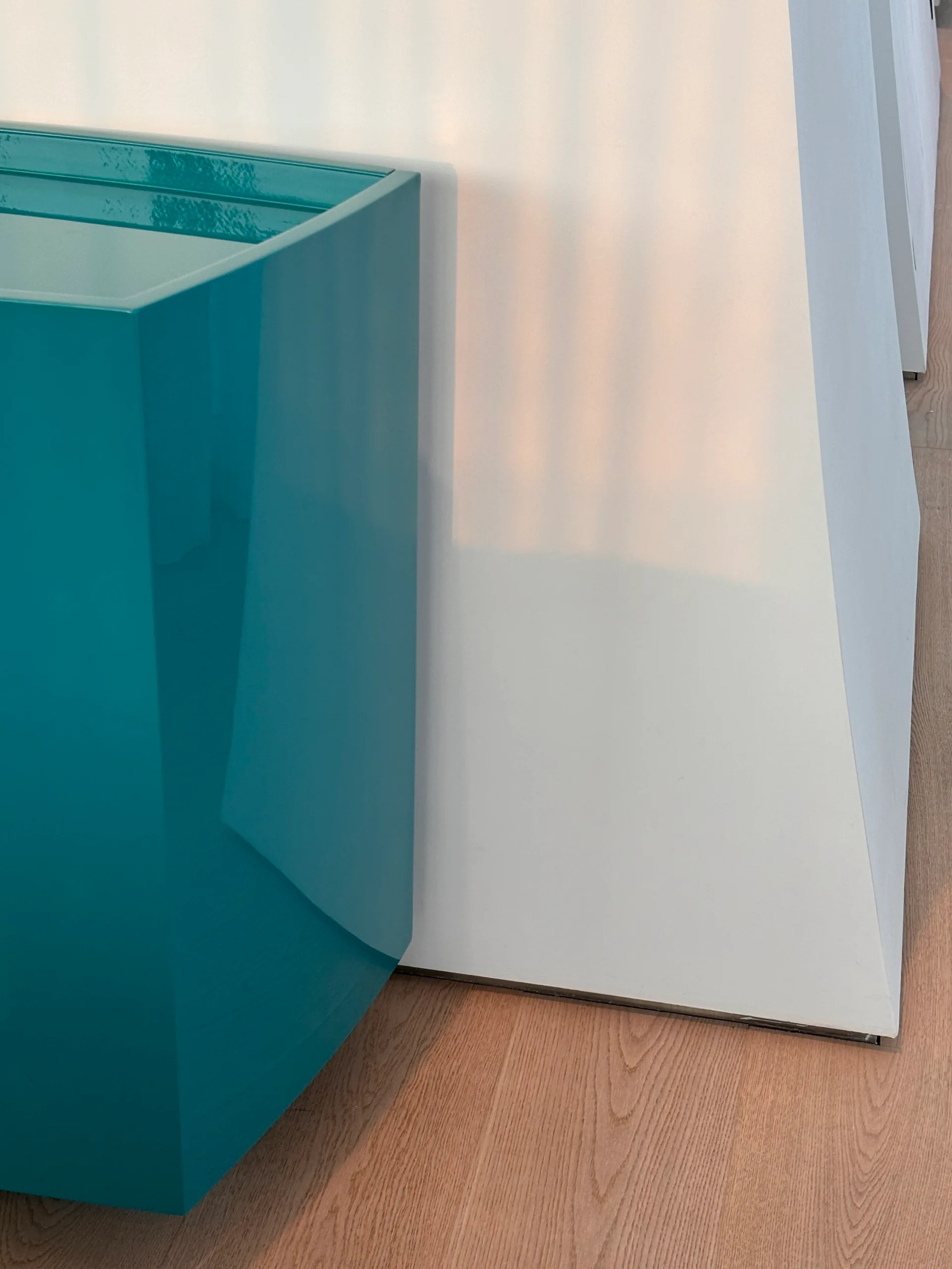 A close-up of a high-gloss teal cabinet positioned against a smooth white wall on light oak wood floors.