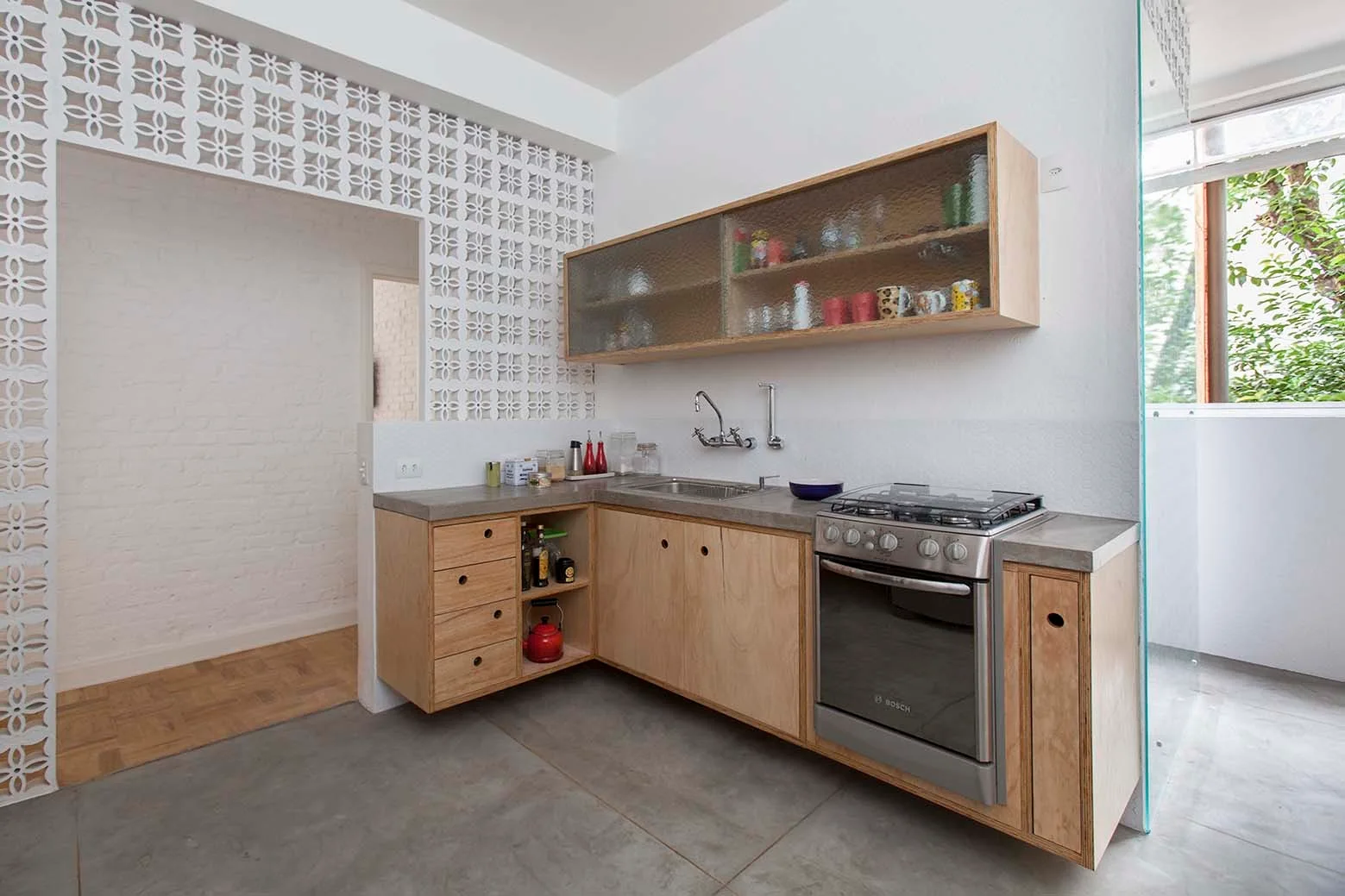 A modern kitchen featuring light plywood cabinetry, a concrete countertop, a stainless steel stove, and a white geometric breeze block partition wall.