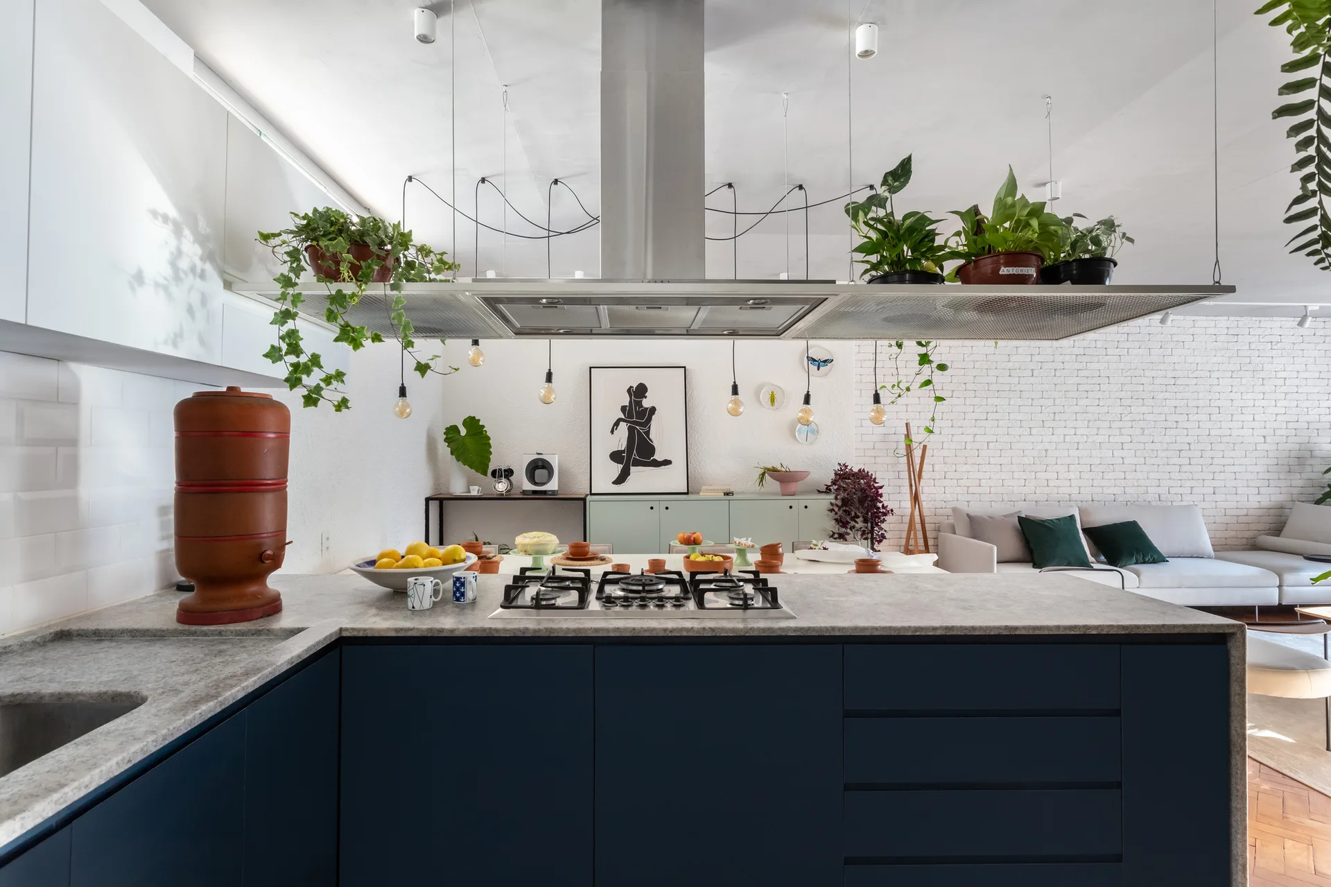 A modern open kitchen features a dark blue island with a stone countertop, a metal range hood topped with potted plants, and a living area in the background.