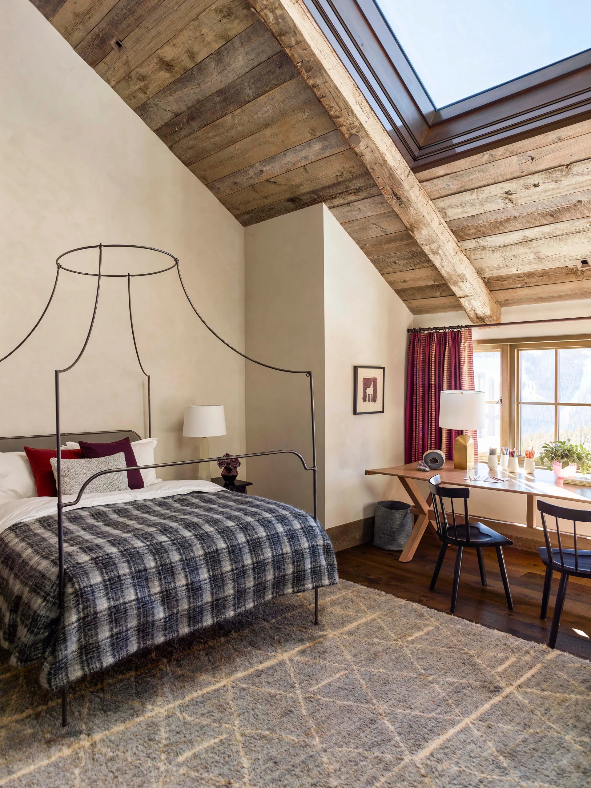 A rustic bedroom with a black metal canopy bed, plaid bedding, a wood-planked sloped ceiling with a skylight, and a desk facing a window.