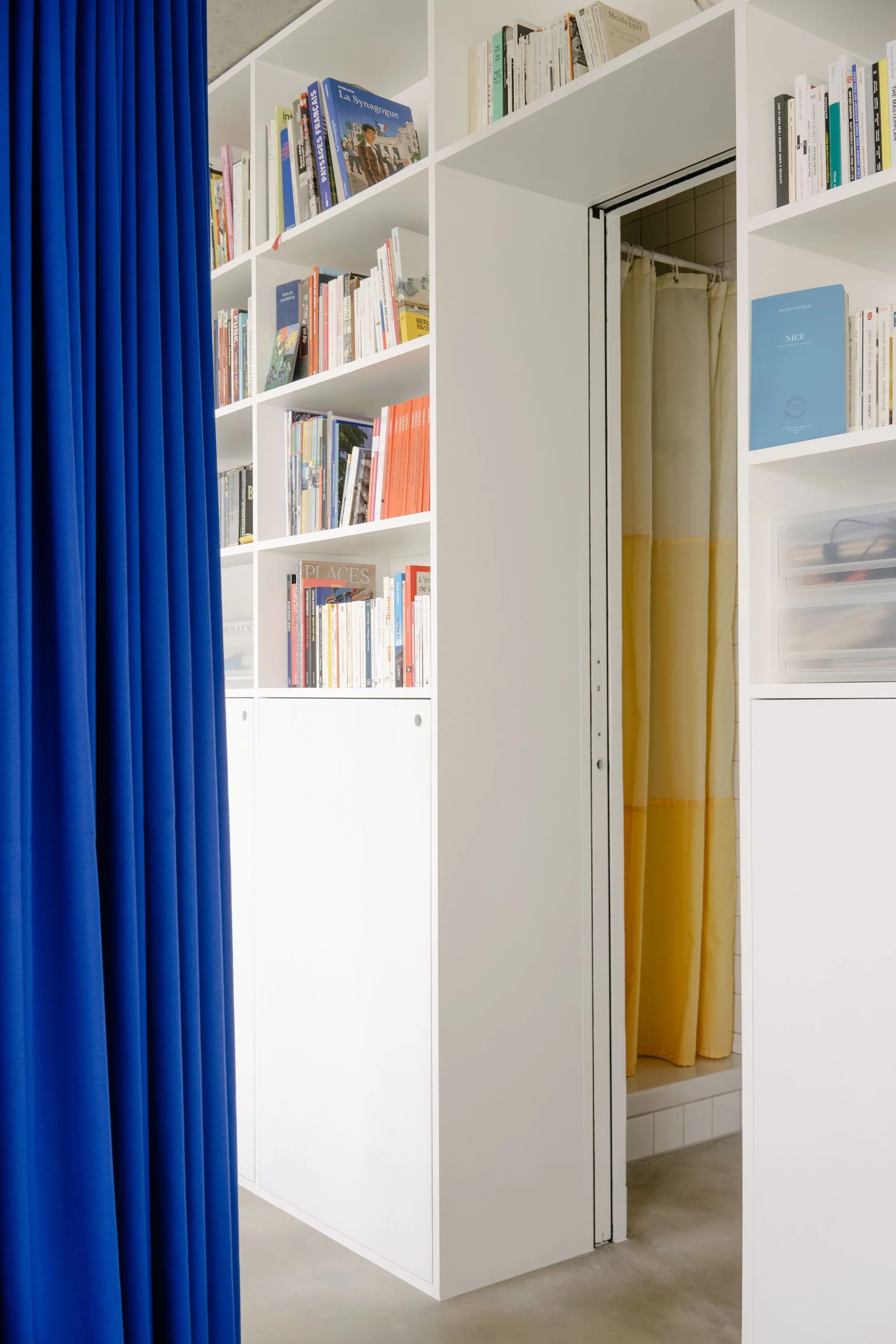 Floor-to-ceiling white shelving units frame an open doorway, with a bright blue curtain in the foreground and a yellow shower curtain visible inside.