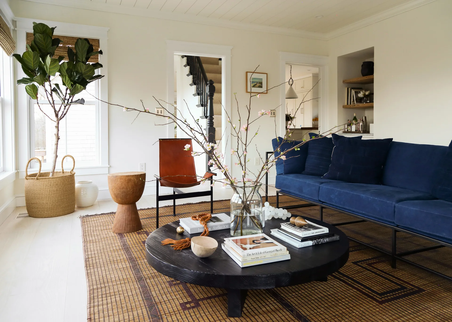 A bright living room features a navy blue sofa and a round black wooden coffee table on a woven rug next to a leather sling chair.