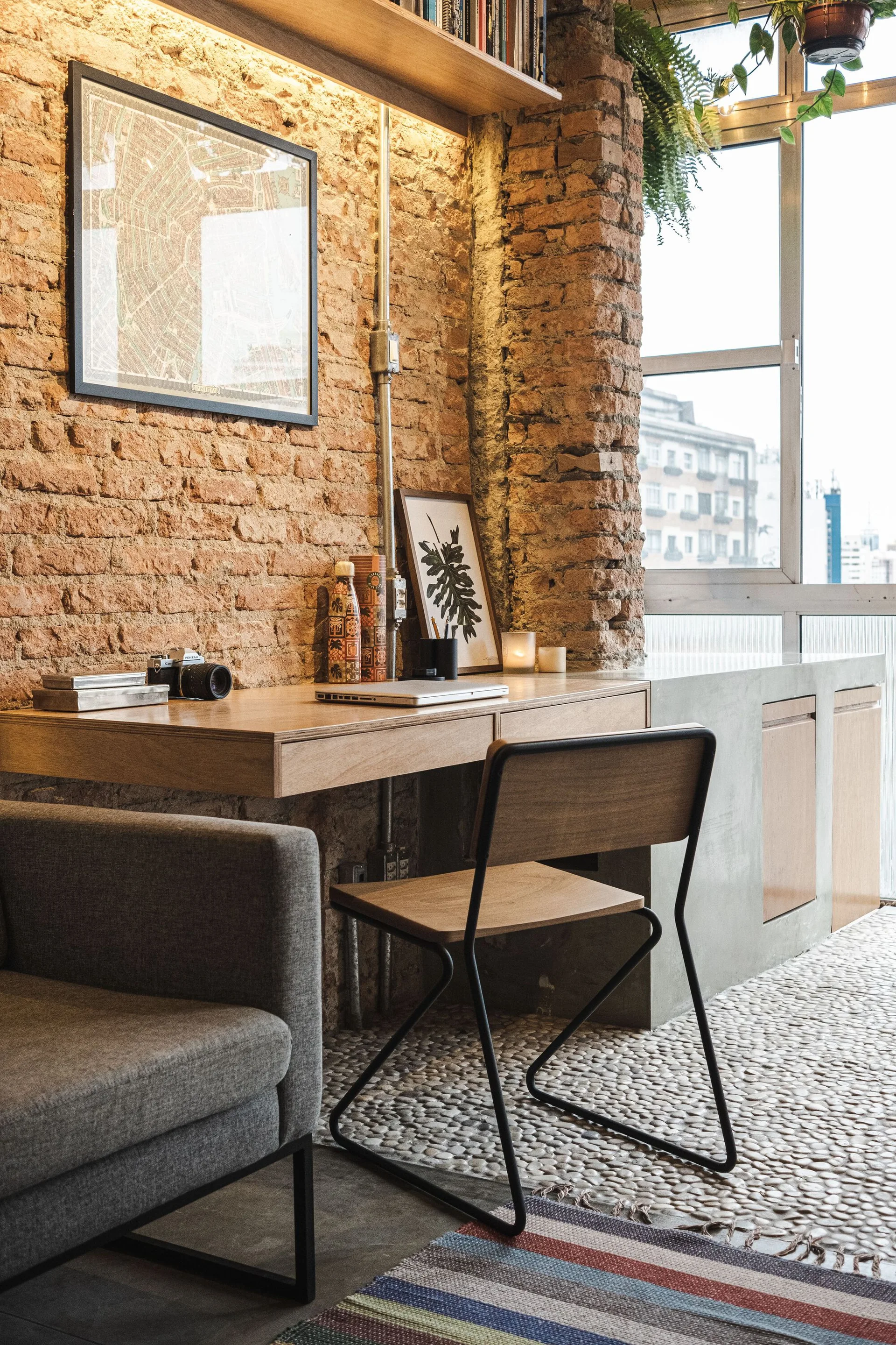 A home office features a wooden built-in desk against an exposed brick wall, a modern wood-and-metal chair, and textured pebble flooring.