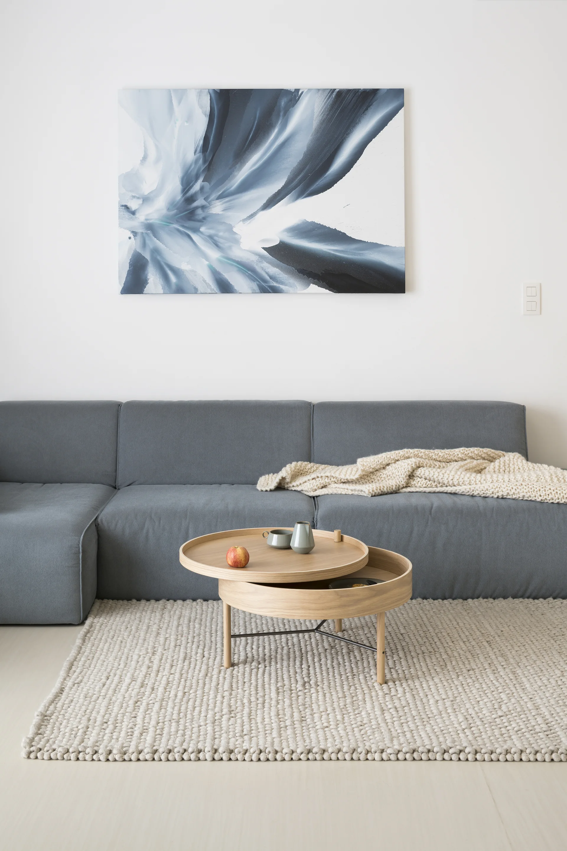 A grey sectional sofa sits on a chunky knit beige rug next to a tiered light wood coffee table beneath a blue abstract painting.