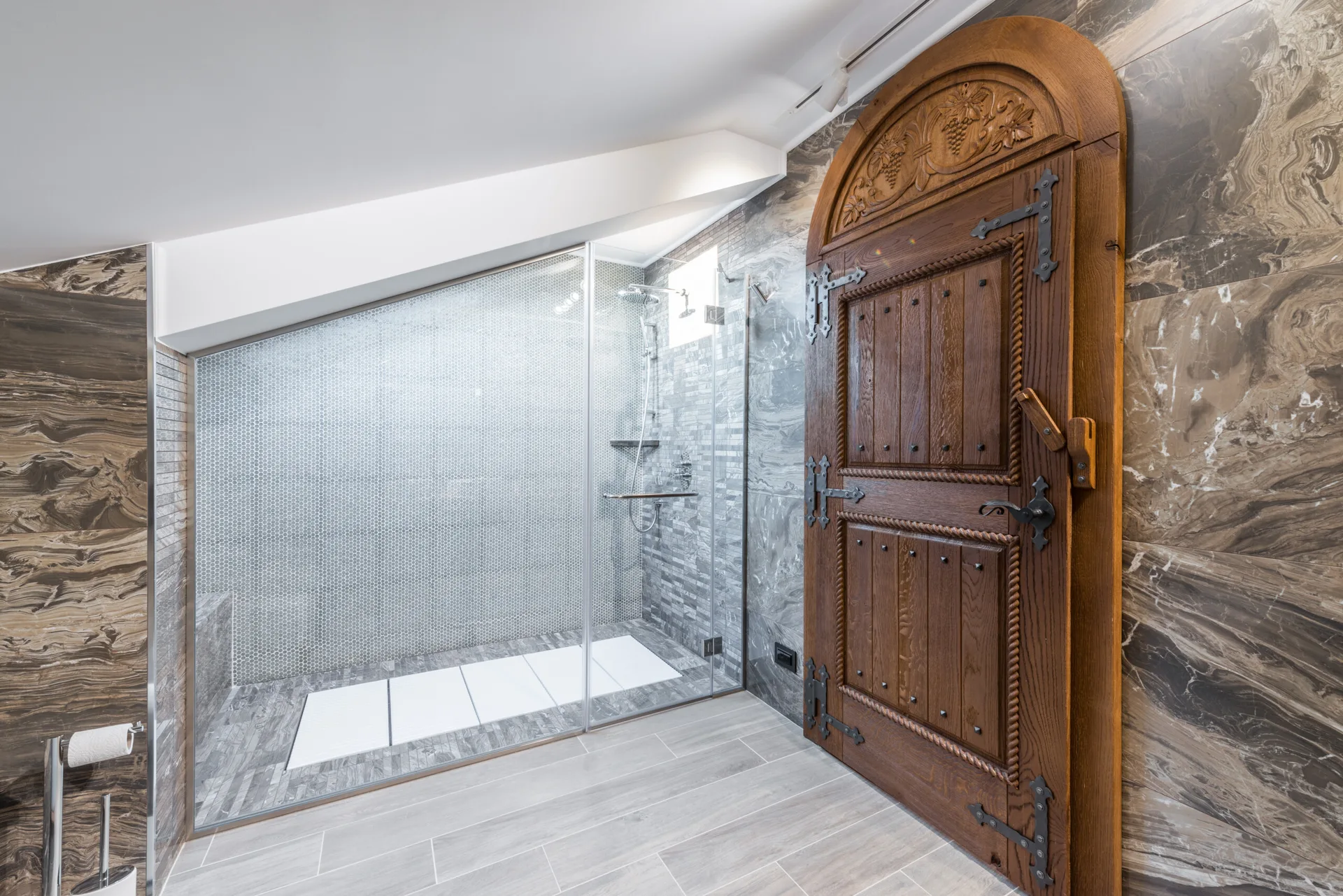 A bathroom featuring an arched wooden door with black metal hinges next to a glass-enclosed walk-in shower with grey mosaic tiling.