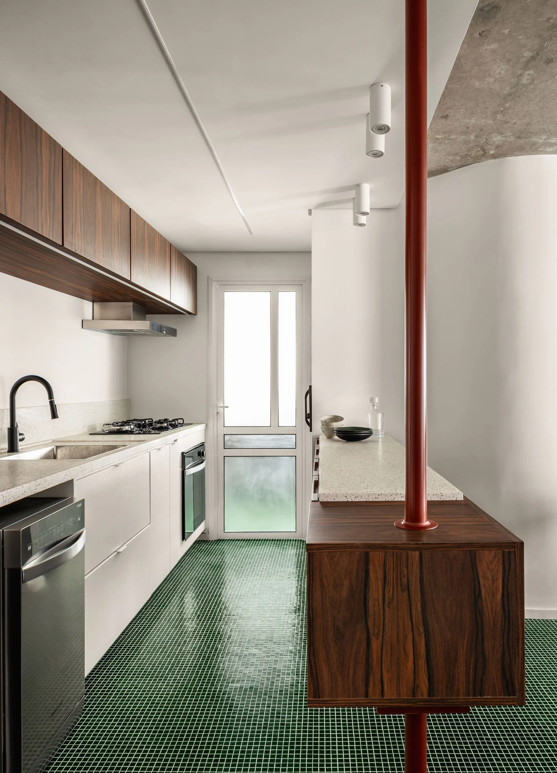 A modern galley kitchen features wood upper cabinets, white lower cabinets, green mosaic floor tiles, and a red metal structural column.