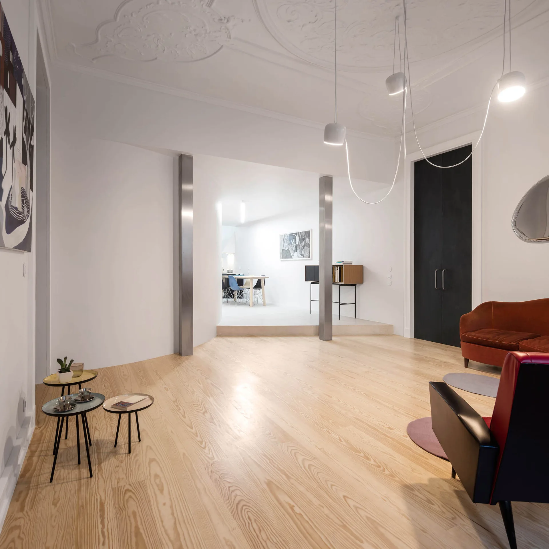 An open living space featuring light wood floors, a decorative white plaster ceiling, a rust-orange sofa, and two metallic support columns.