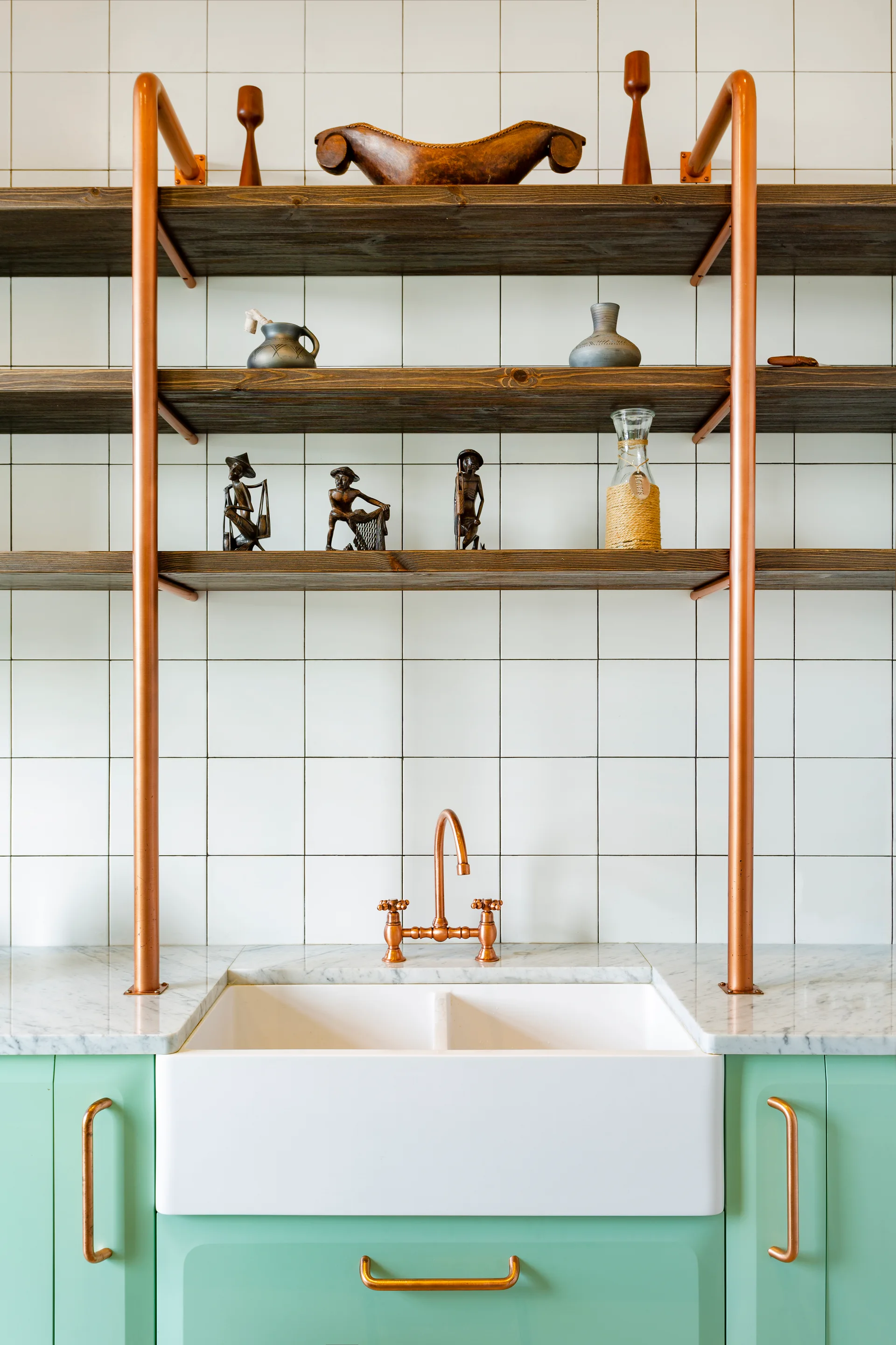 A white farmhouse sink with a copper faucet sits below open wooden shelves supported by copper piping against a white tiled wall.