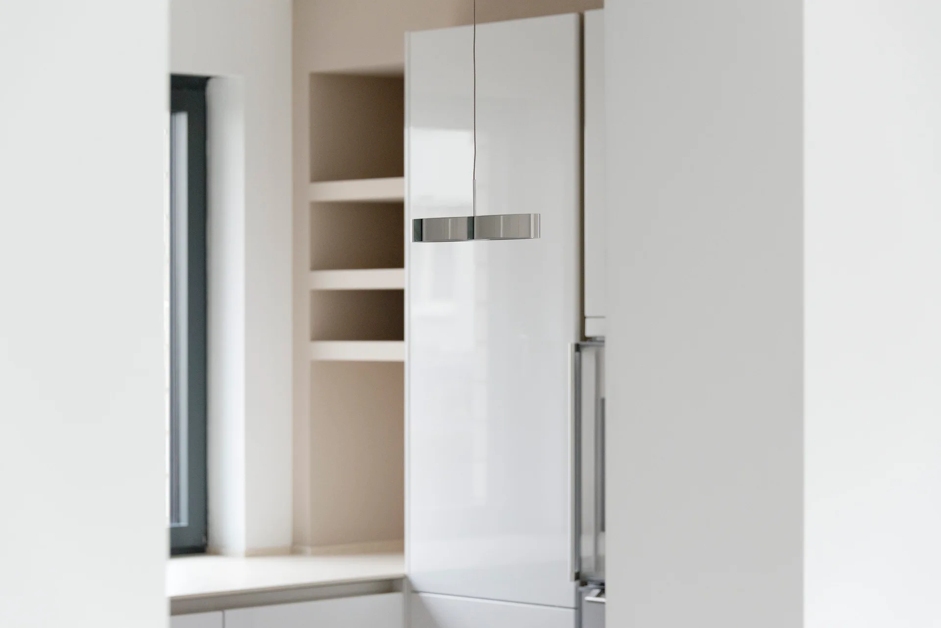 A minimalist kitchen view featuring glossy white cabinetry, a recessed beige shelving niche, and a slim metallic pendant light.