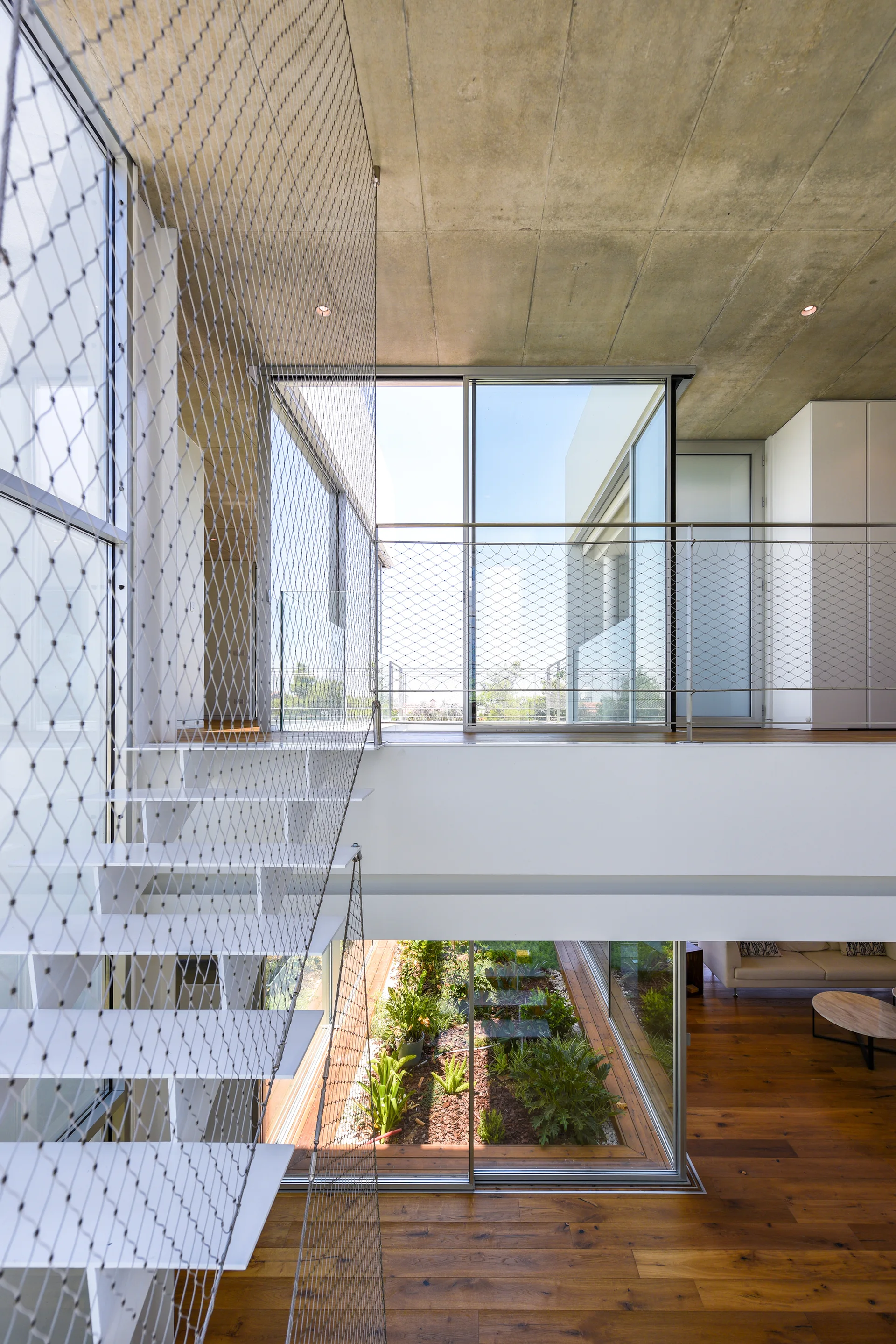 A white metal floating staircase with a wire mesh safety railing overlooks an indoor courtyard garden inside a modern home.