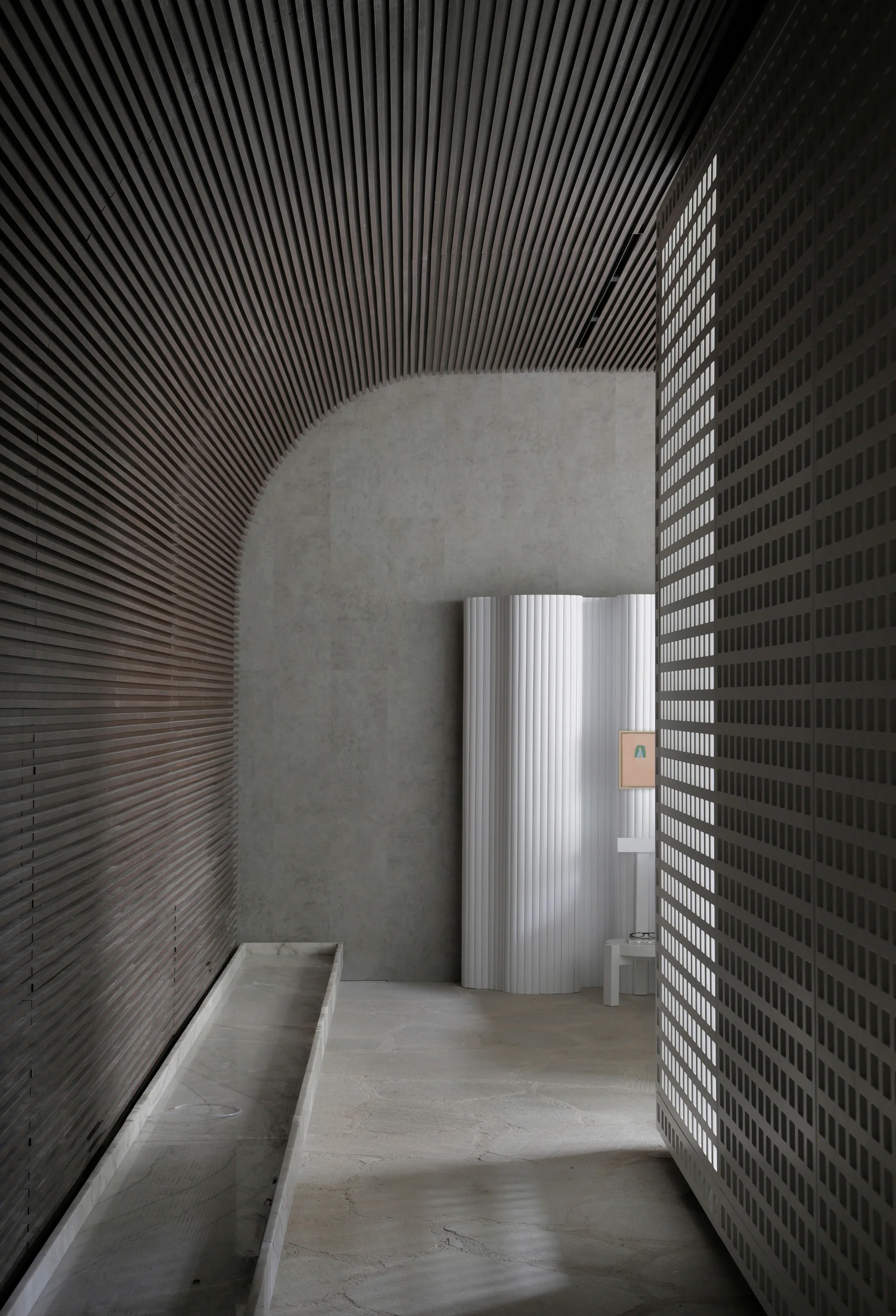 A minimalist hallway featuring a curved dark wood slatted ceiling, a textured stone floor, a white fluted partition, and a grid-patterned screen.