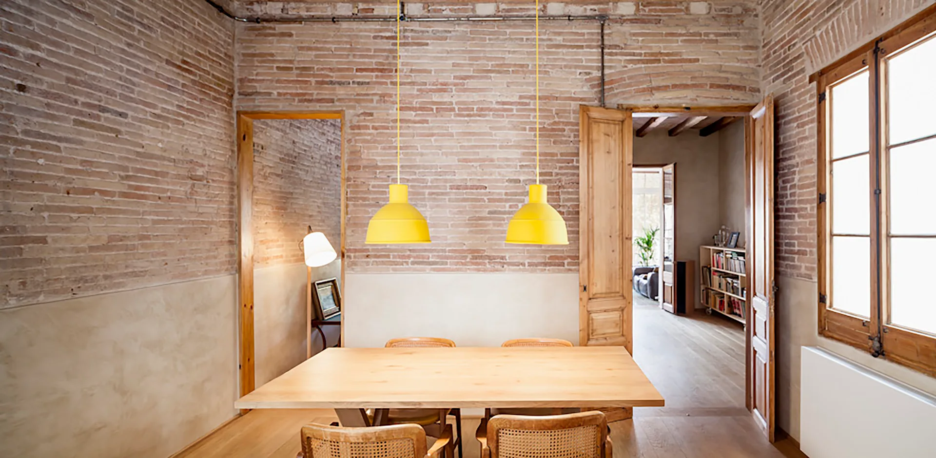 A rustic dining room features a light wood table, woven cane chairs, two bright yellow pendant lights, and exposed brick walls with plaster wainscoting.