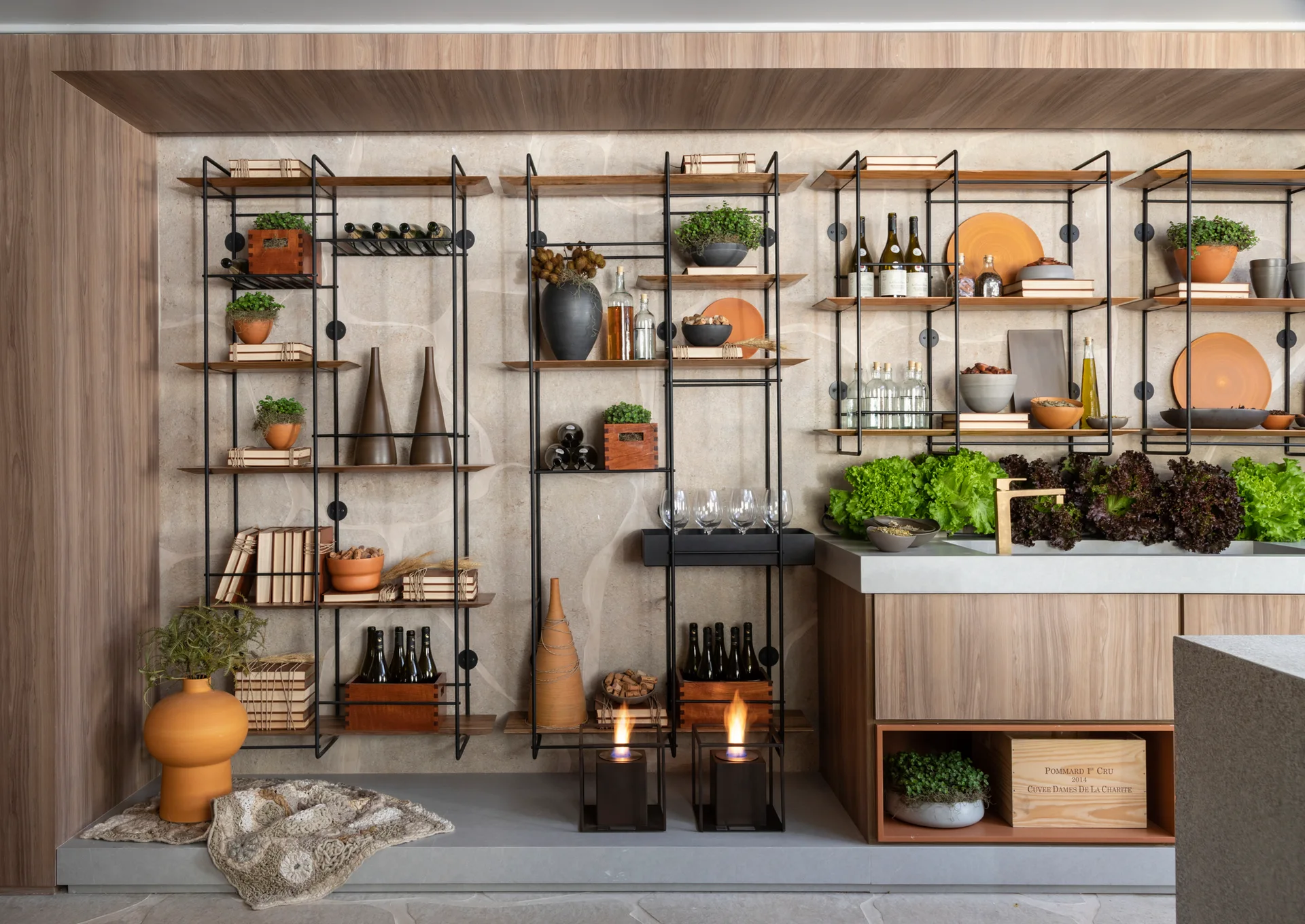 Open black metal shelving filled with wine bottles and ceramic tableware stands against a beige stone wall next to wood-paneled kitchen cabinets.