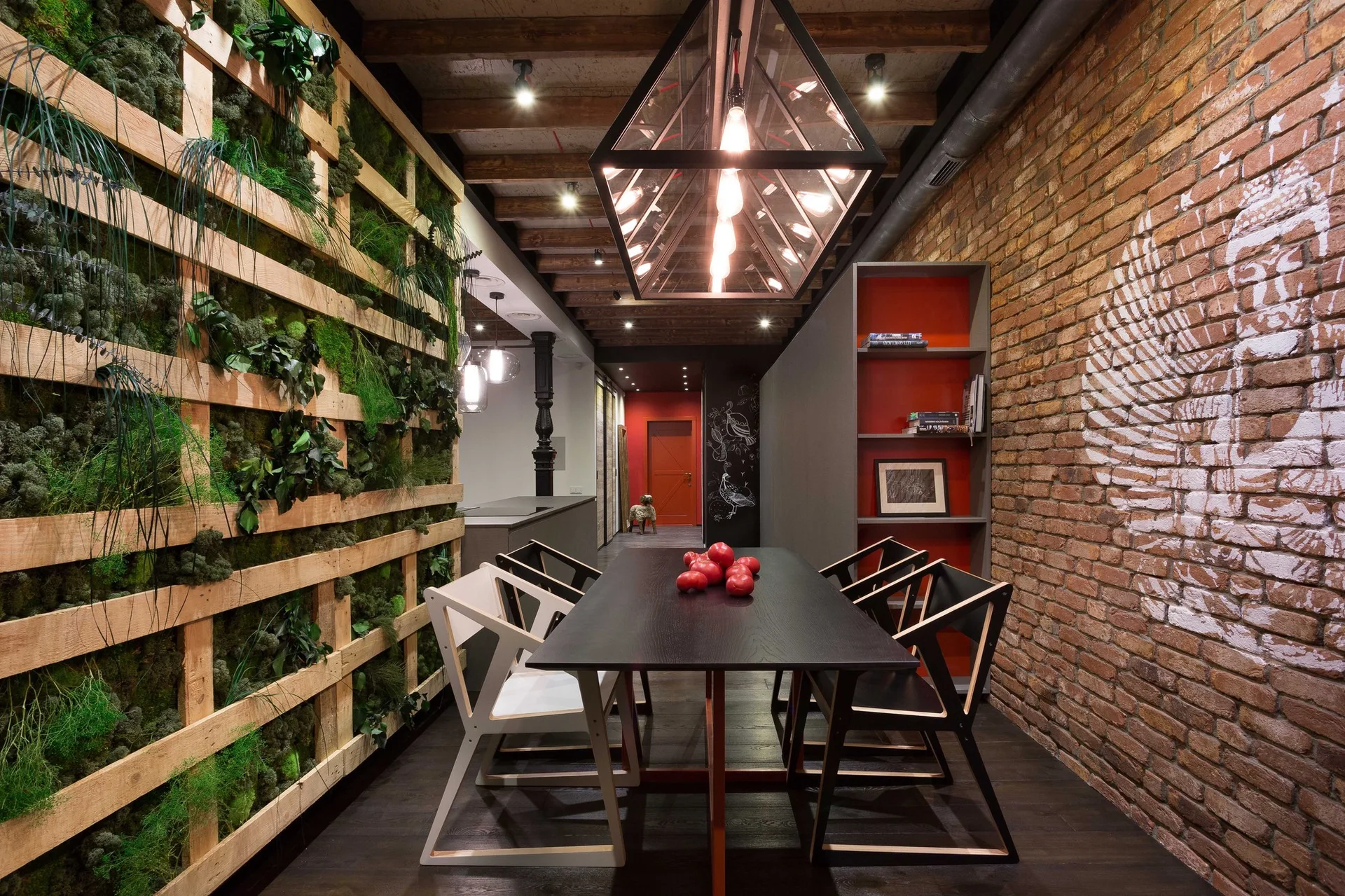 An industrial dining room features a long black table under a geometric pendant light, flanked by a lush green living wall and an exposed brick wall.