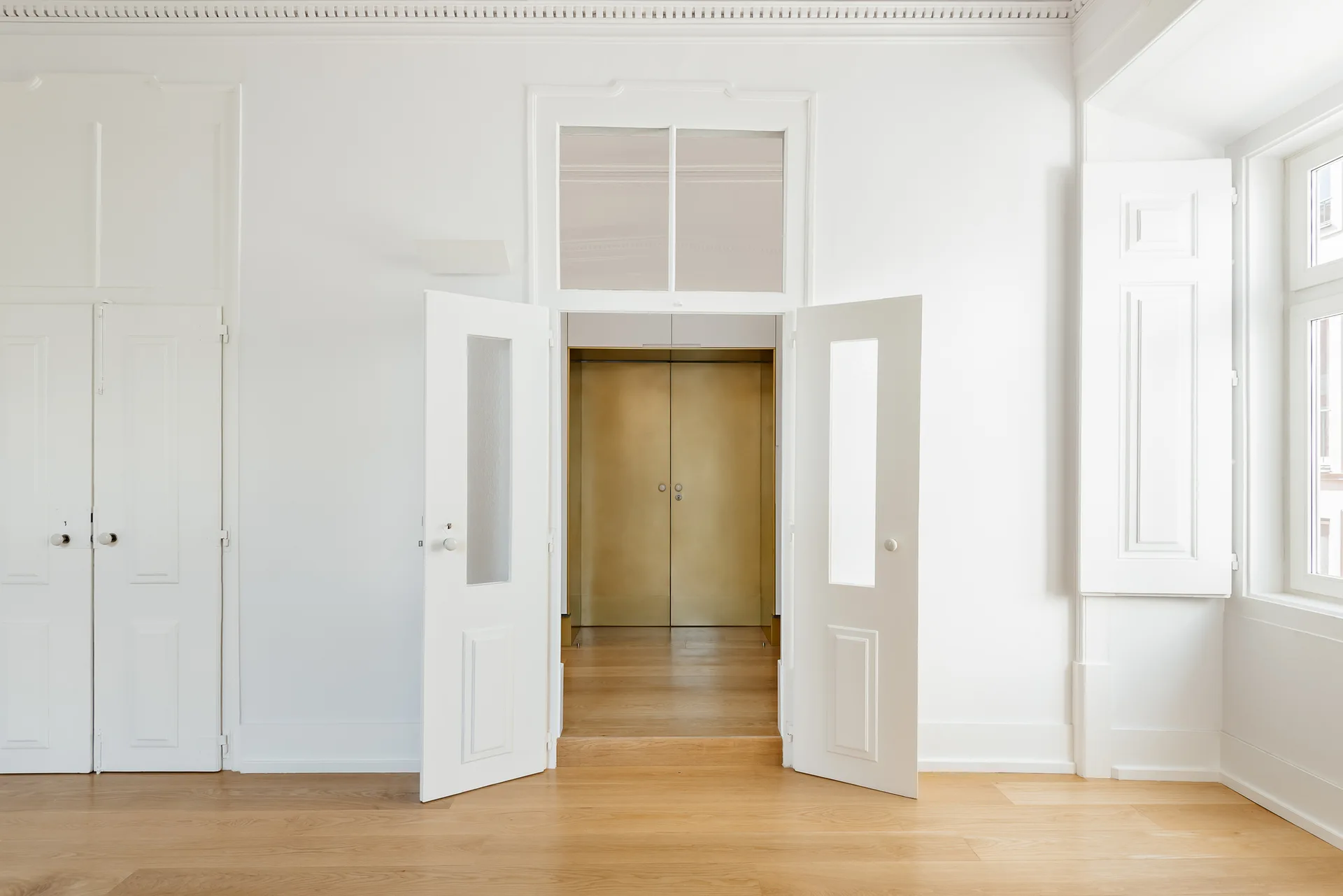 A white double door with window glass panels opens to reveal a set of metallic gold doors at the end of a short hallway.