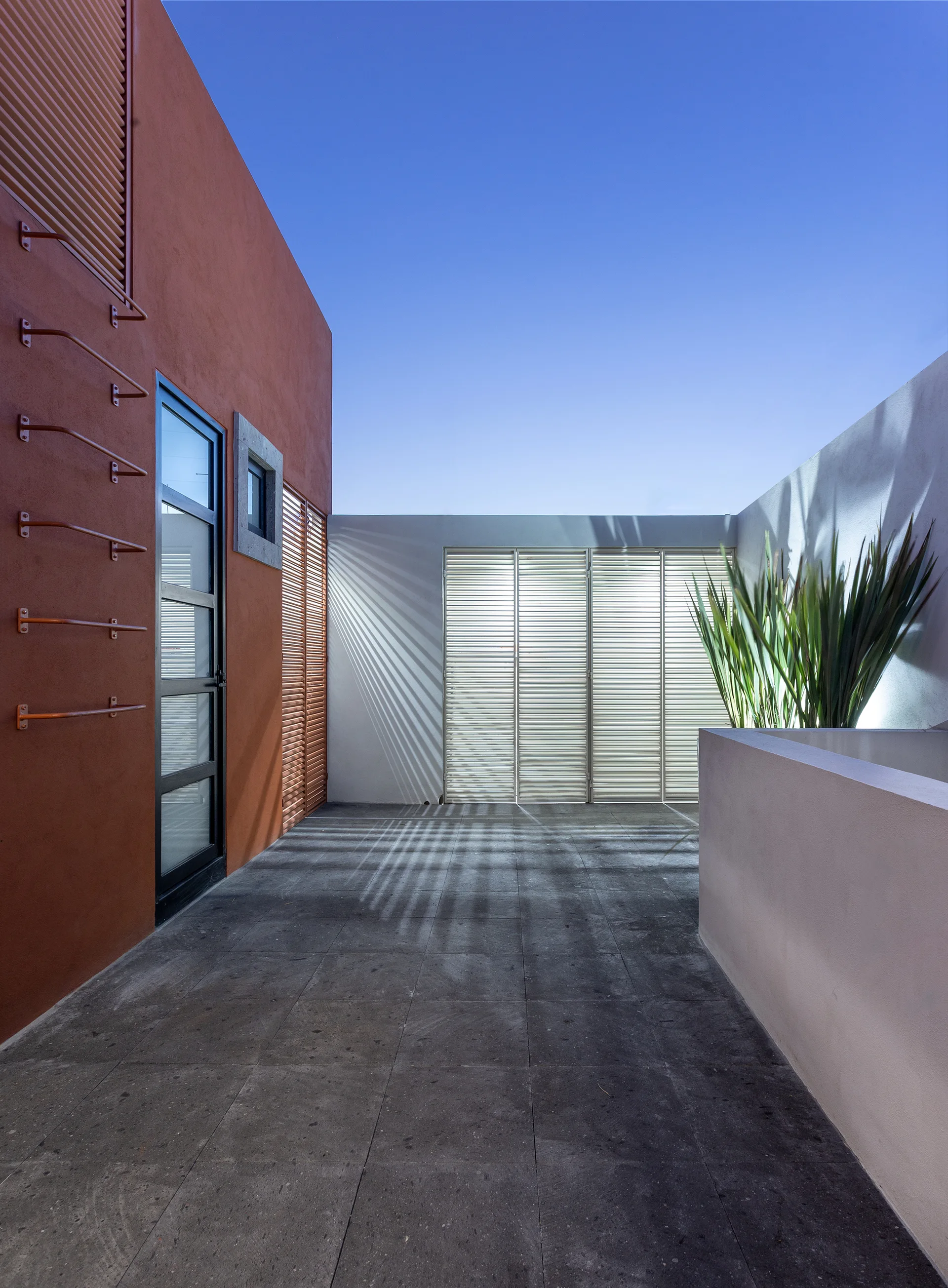 An outdoor rooftop terrace features a bold terracotta stucco wall, dark grey stone floor tiles, and a white wall catching shadows from a slatted screen.