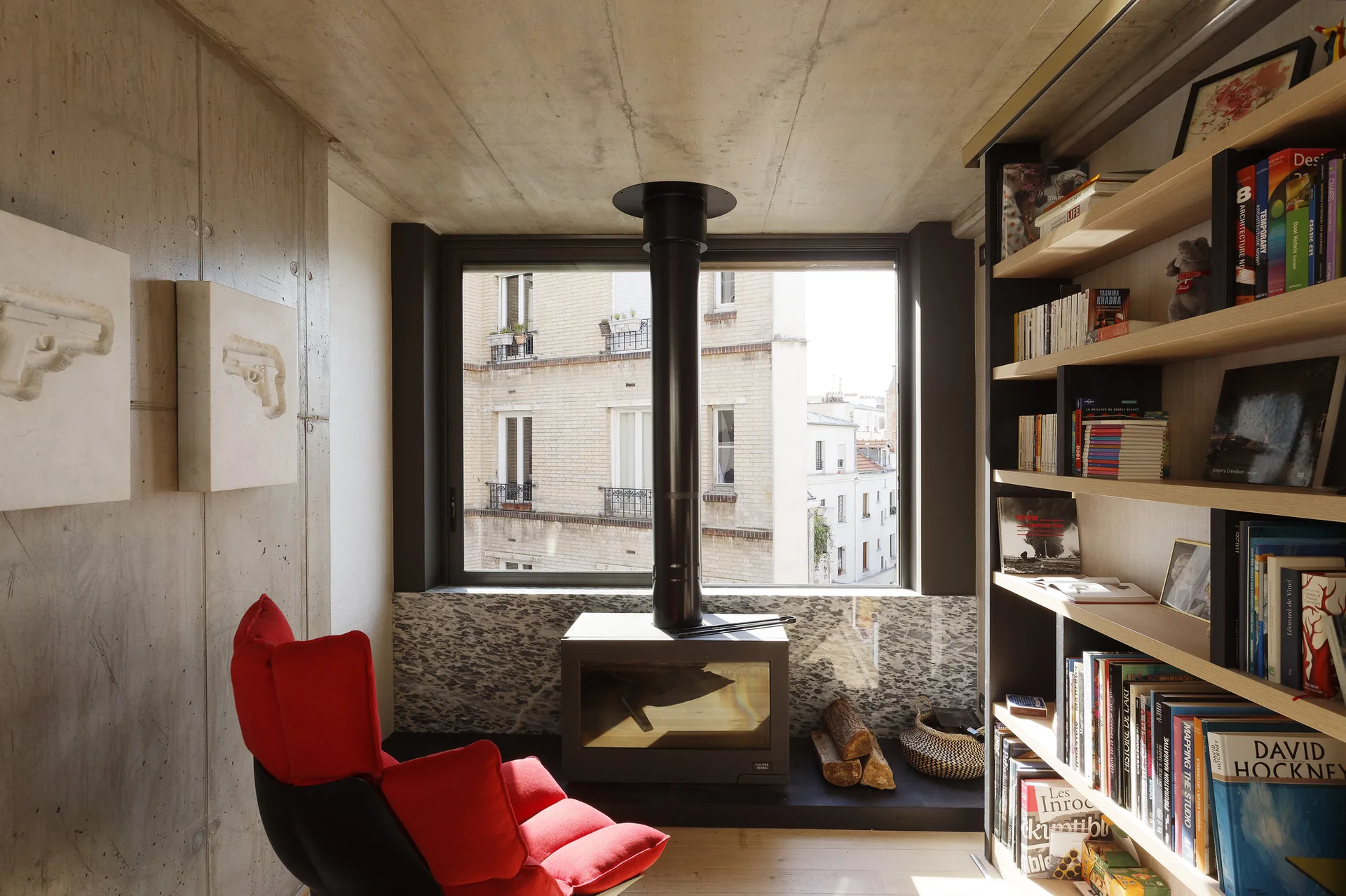 A minimalist room featuring raw concrete walls, a central black wood stove by the window, a bright red lounge chair, and wood bookshelves.