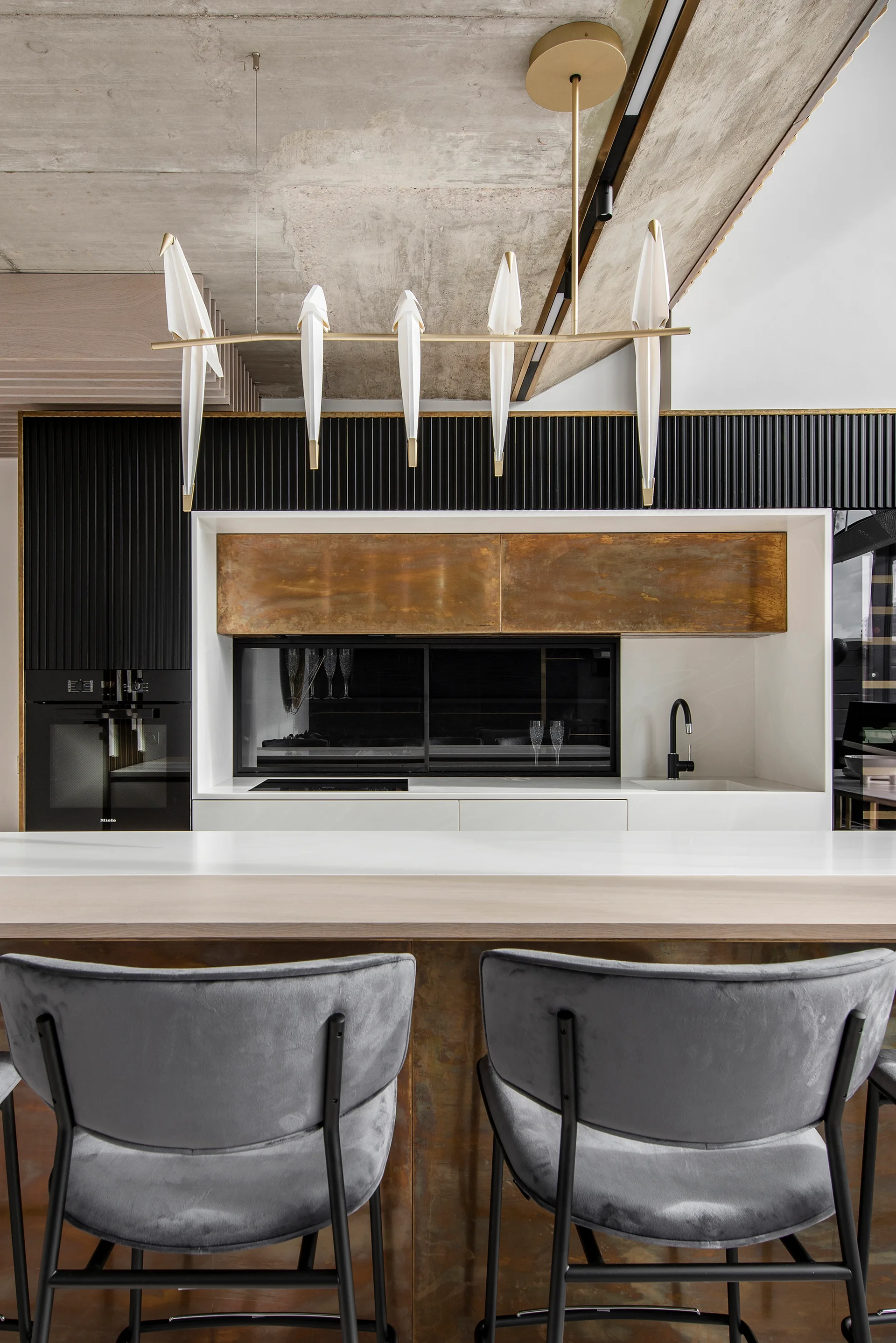 A modern kitchen features gray upholstered bar stools at an island beneath a bird-shaped pendant light, with black fluted cabinets and a concrete ceiling.