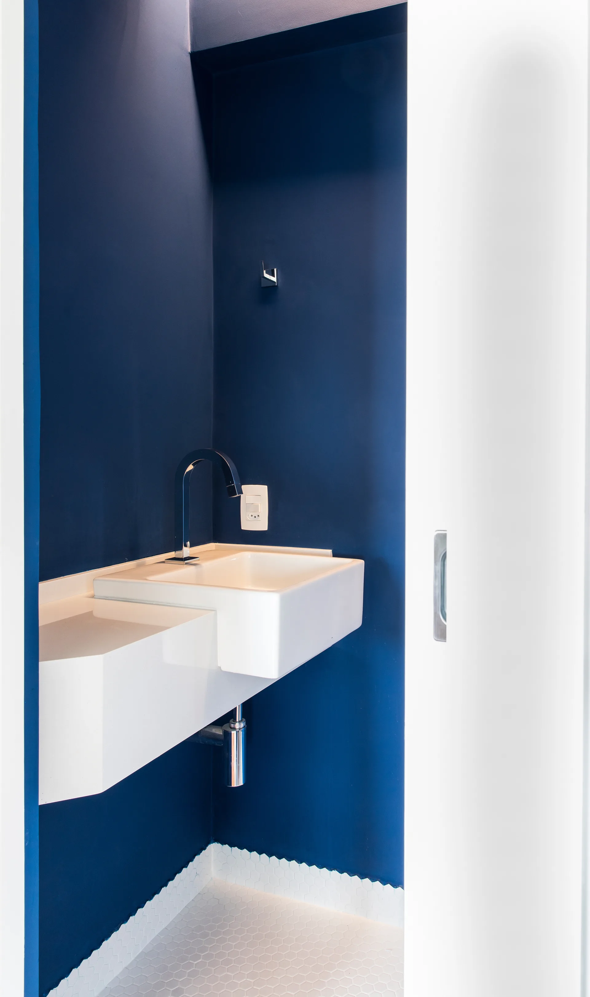 A small powder room features a white wall-mounted sink against deep navy blue walls and white hexagonal tiled floors.