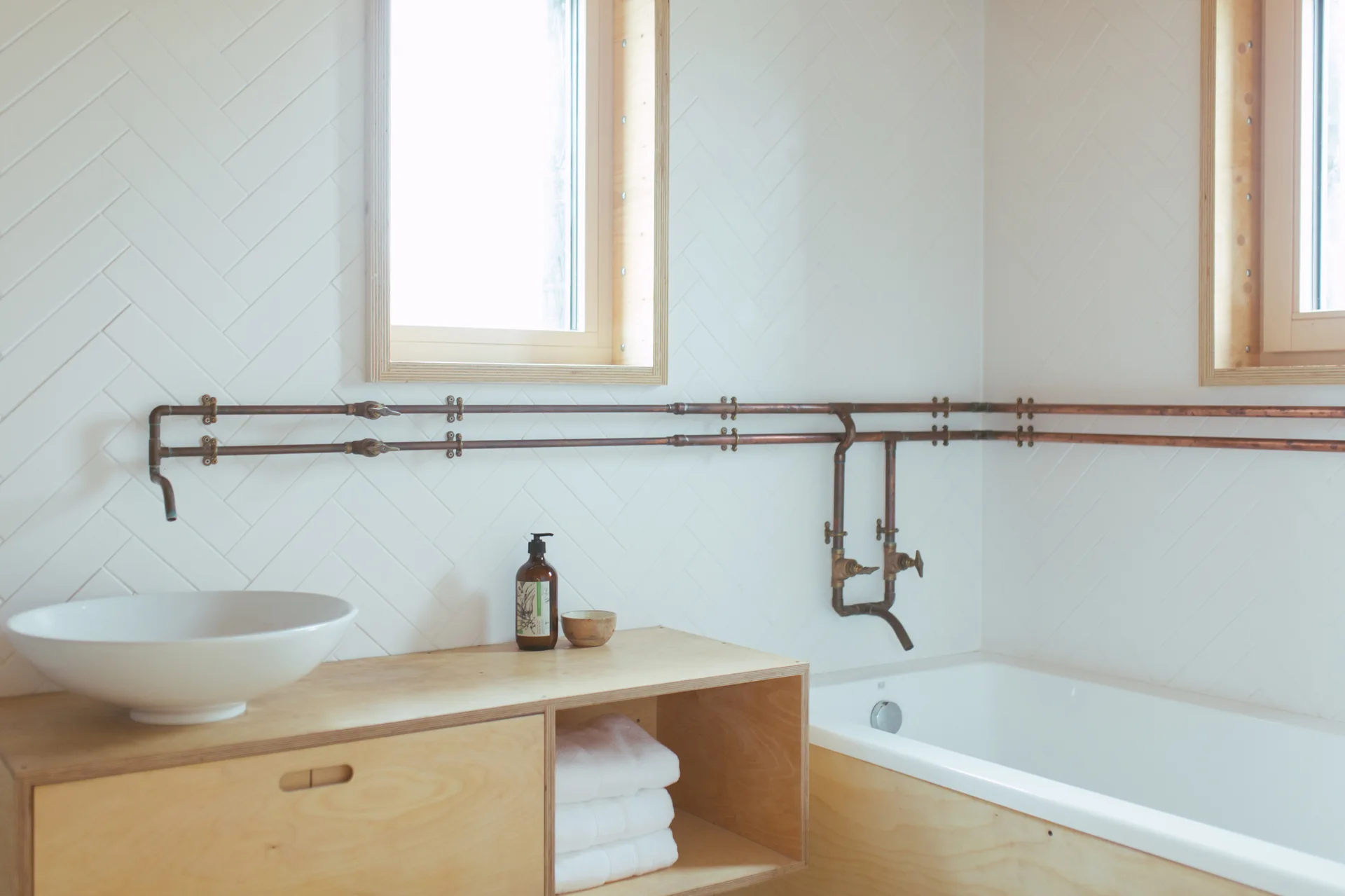 A bright bathroom features a light plywood vanity with a white vessel sink next to a bathtub under exposed copper piping on herringbone tile walls.
