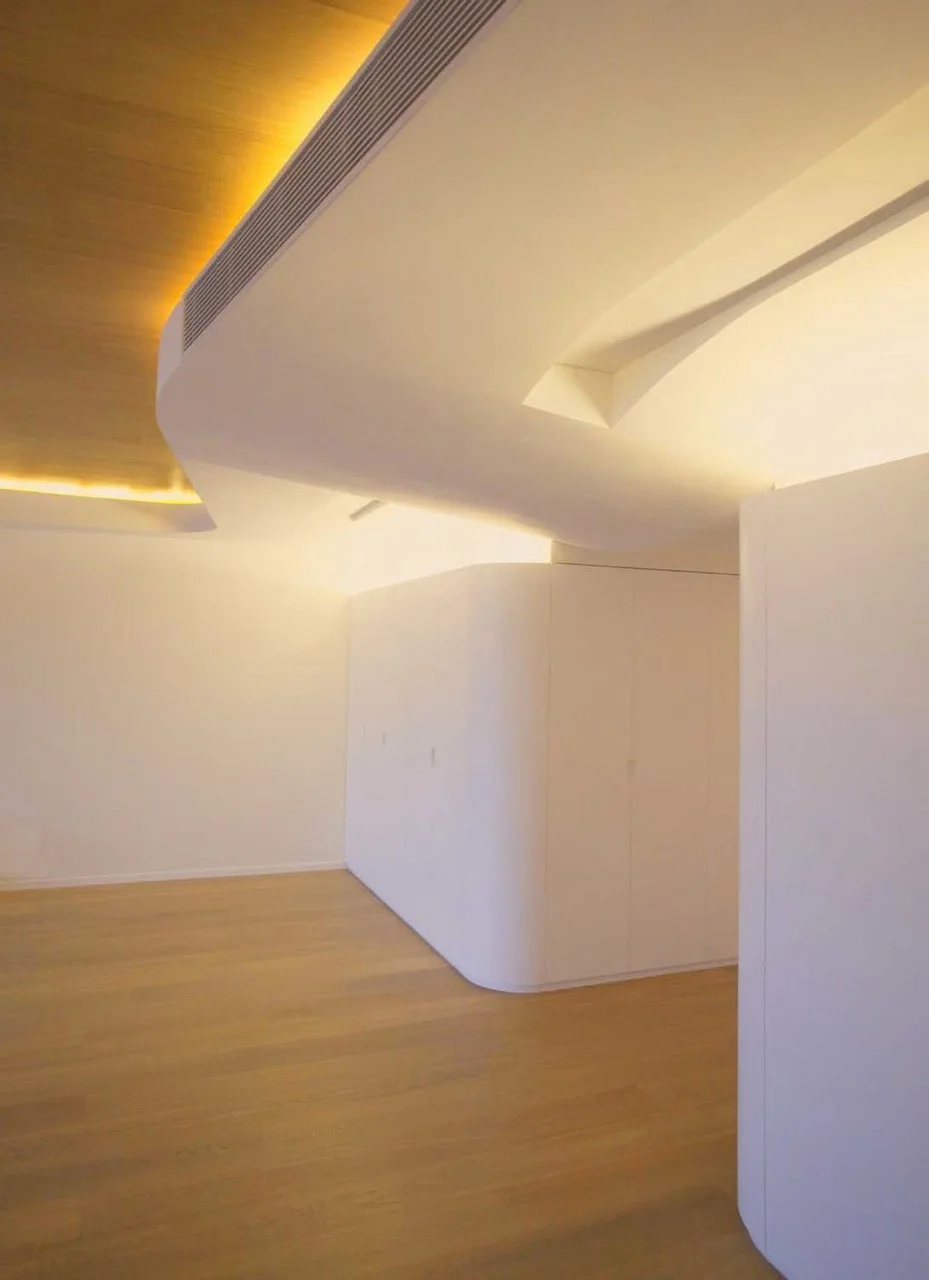 A modern minimalist interior featuring curved white built-in cabinetry, light wood floors, and a curved ceiling soffit with integrated warm cove lighting.