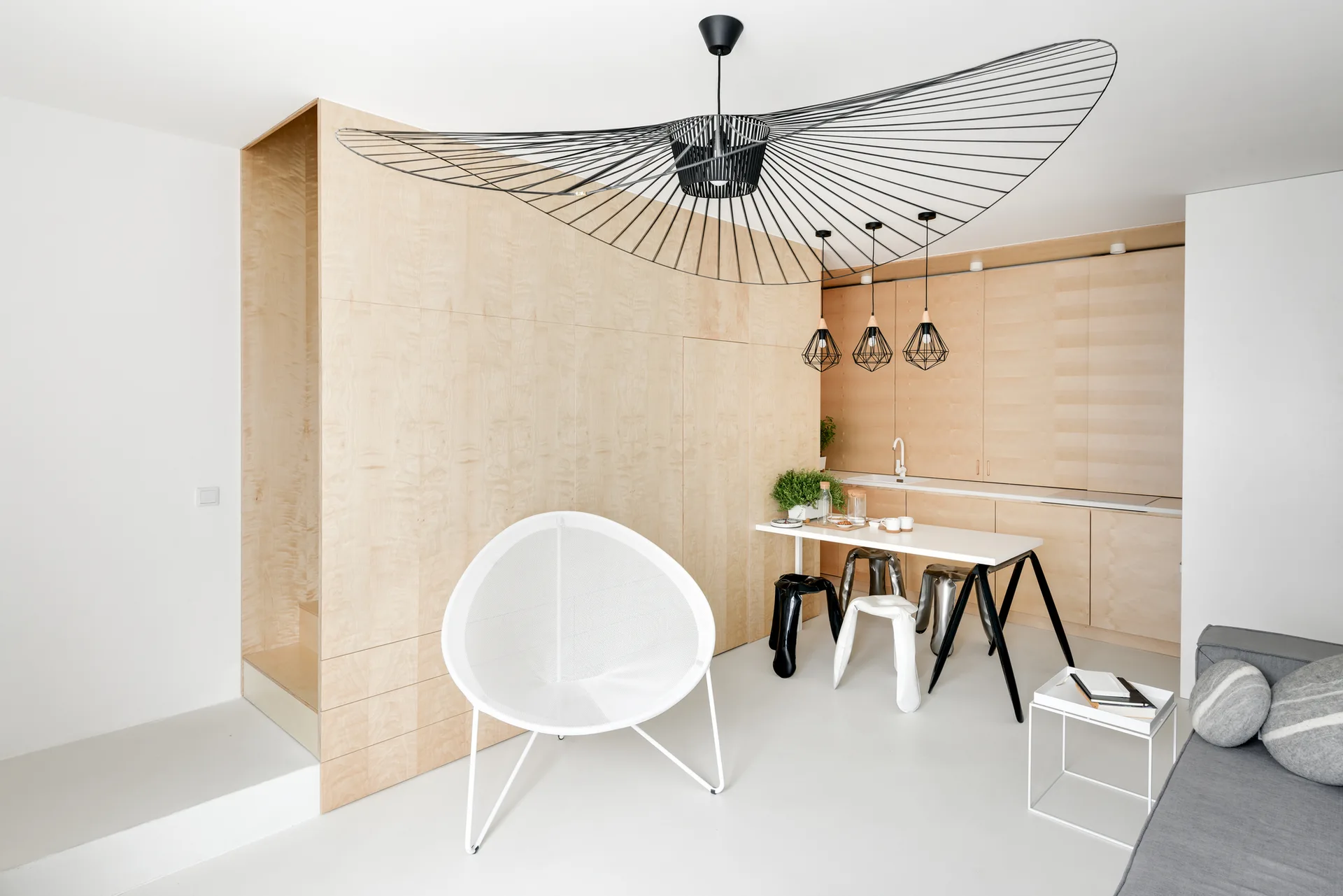 A minimalist studio features light wood wall paneling, an oversized black sculptural pendant light, a white circular mesh chair, and a small dining table.
