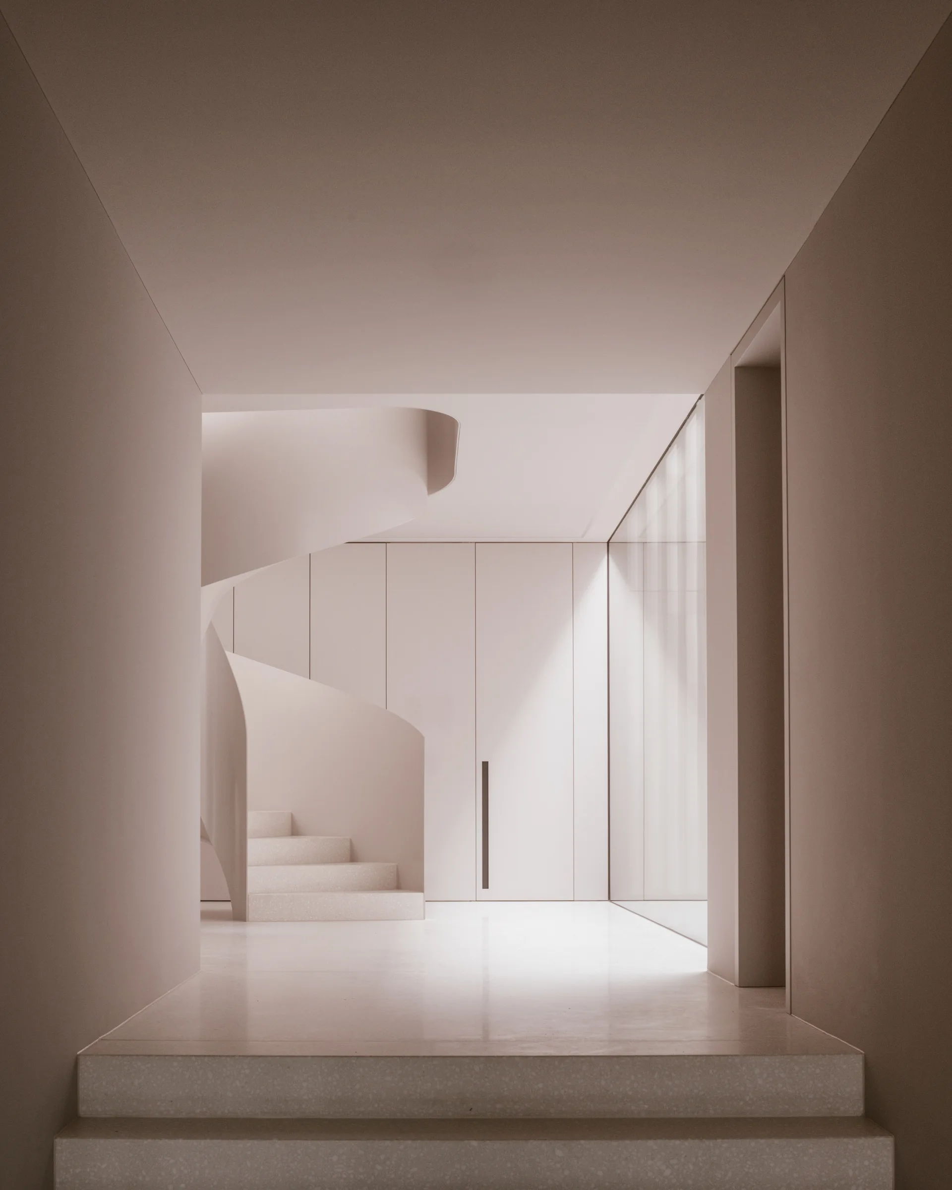 A minimalist beige hallway features a curved sculptural staircase, seamless built-in closet panels, and a large floor-to-ceiling glass wall.