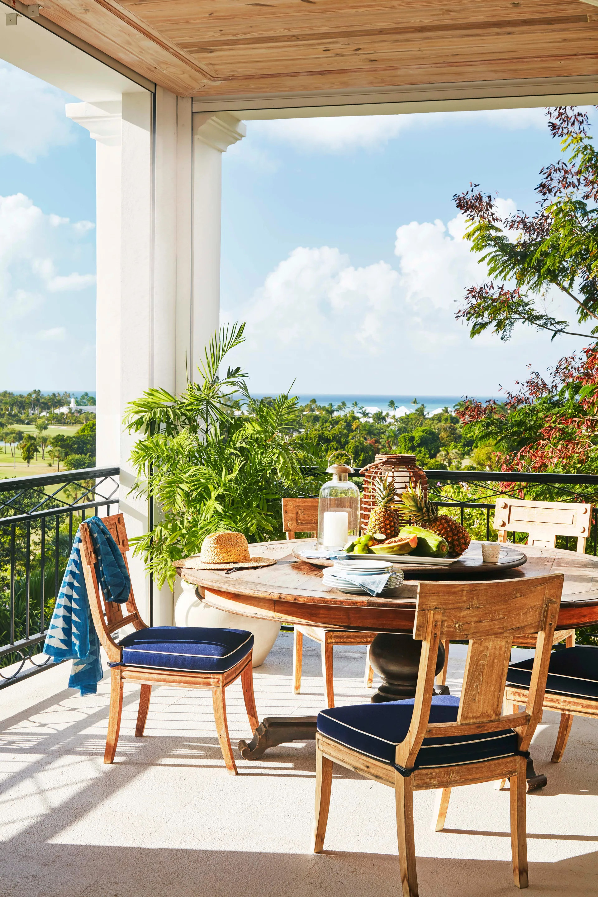 A covered porch features a round wooden dining table and matching chairs with navy blue cushions, overlooking a lush green landscape and the ocean.