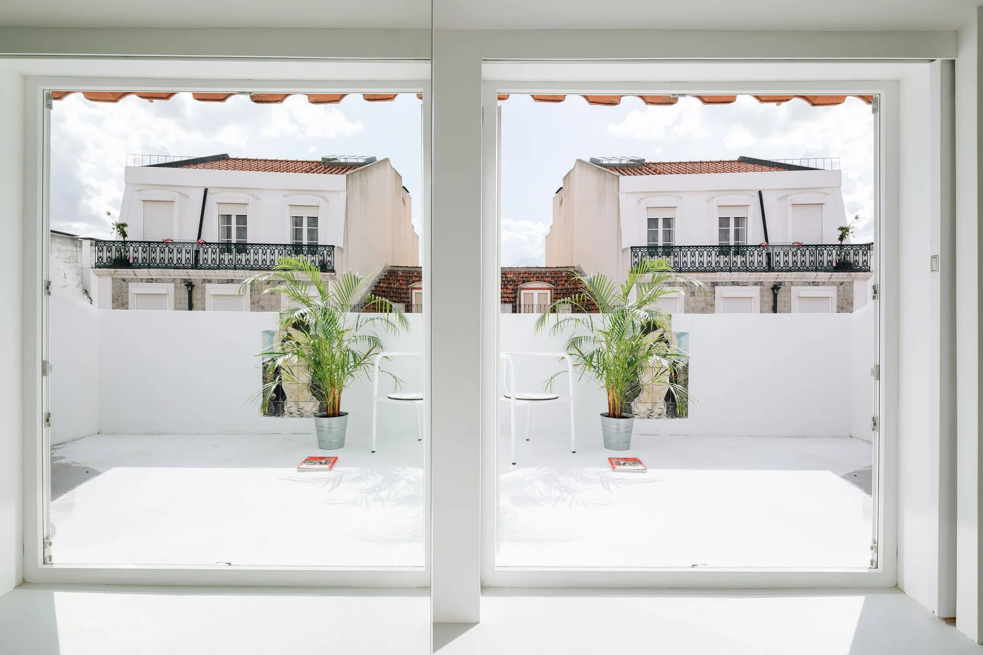 A bright white minimalist terrace with potted palm plants and white chairs, viewed through a large window next to a mirrored wall panel.