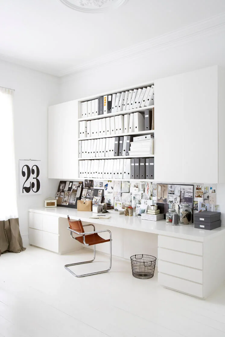 A bright white home office featuring a long built-in desk, a brown leather cantilever chair, and overhead shelves filled with organized binders.