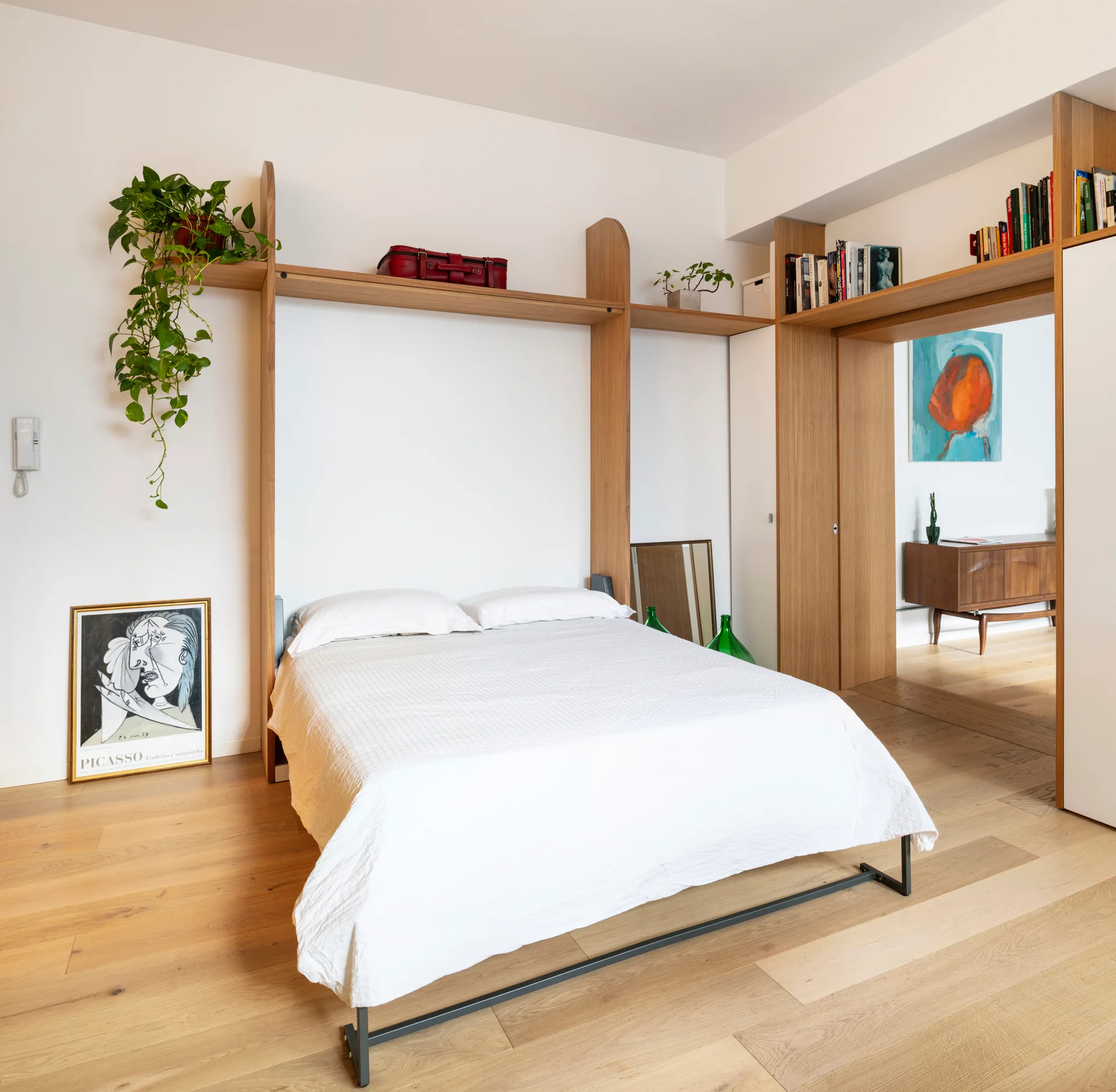 A bright bedroom featuring a white bed framed by light wood wall shelving, next to a wide wooden doorway opening to another room.