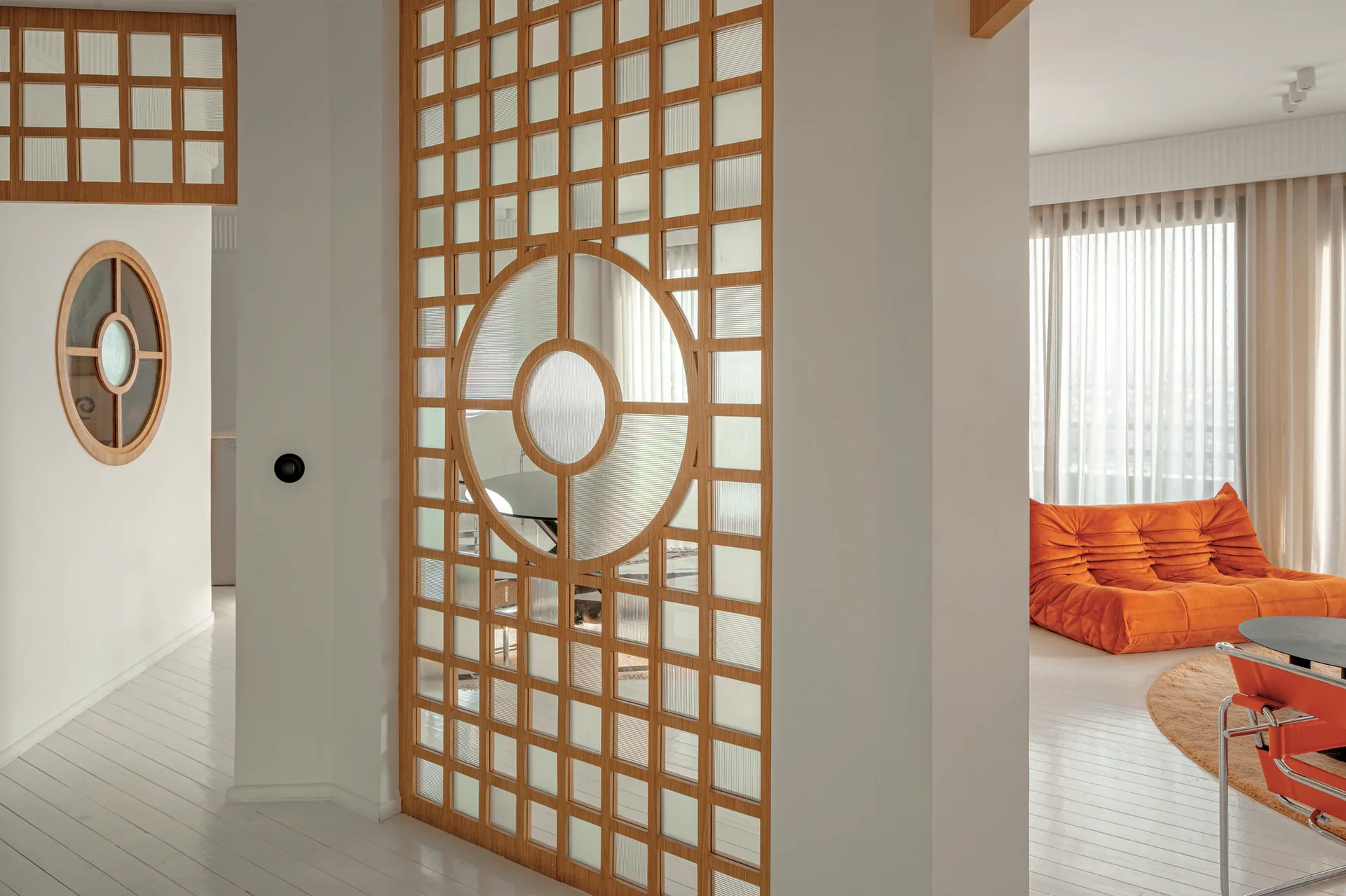 A geometric wood and reeded glass partition divides a bright space featuring white plank floors and a low-slung orange sofa.