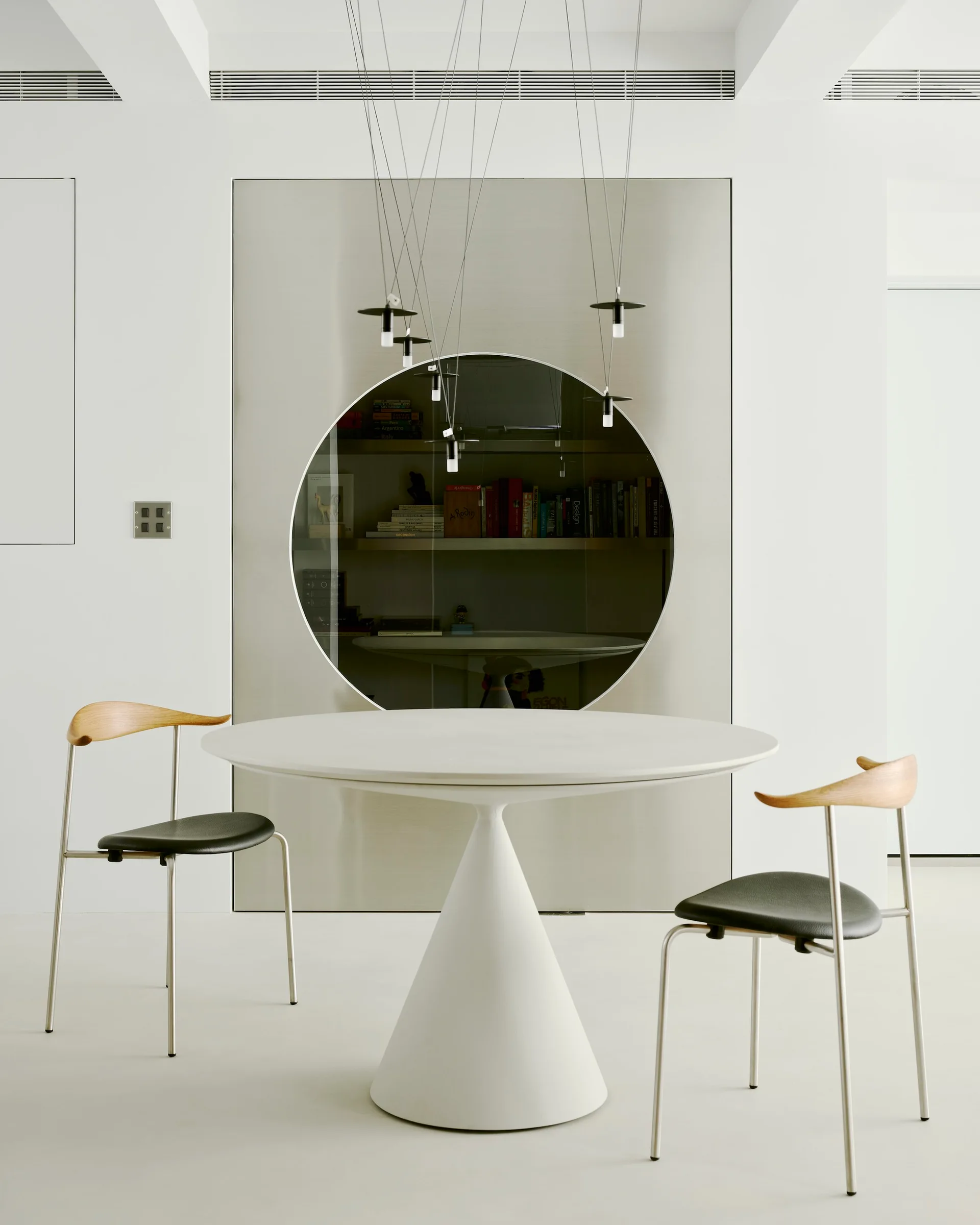 A white pedestal dining table and two modern chairs sit in front of a metallic accent wall panel with a large circular cutout showing bookshelves.