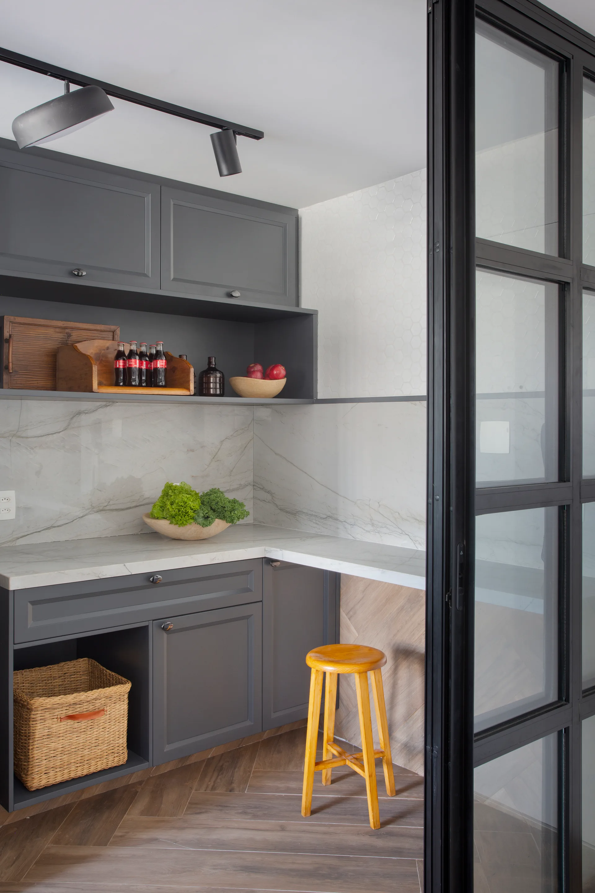 A kitchen corner features dark grey cabinetry, a marble backsplash, herringbone floors, a wooden stool, and a black-framed glass door.