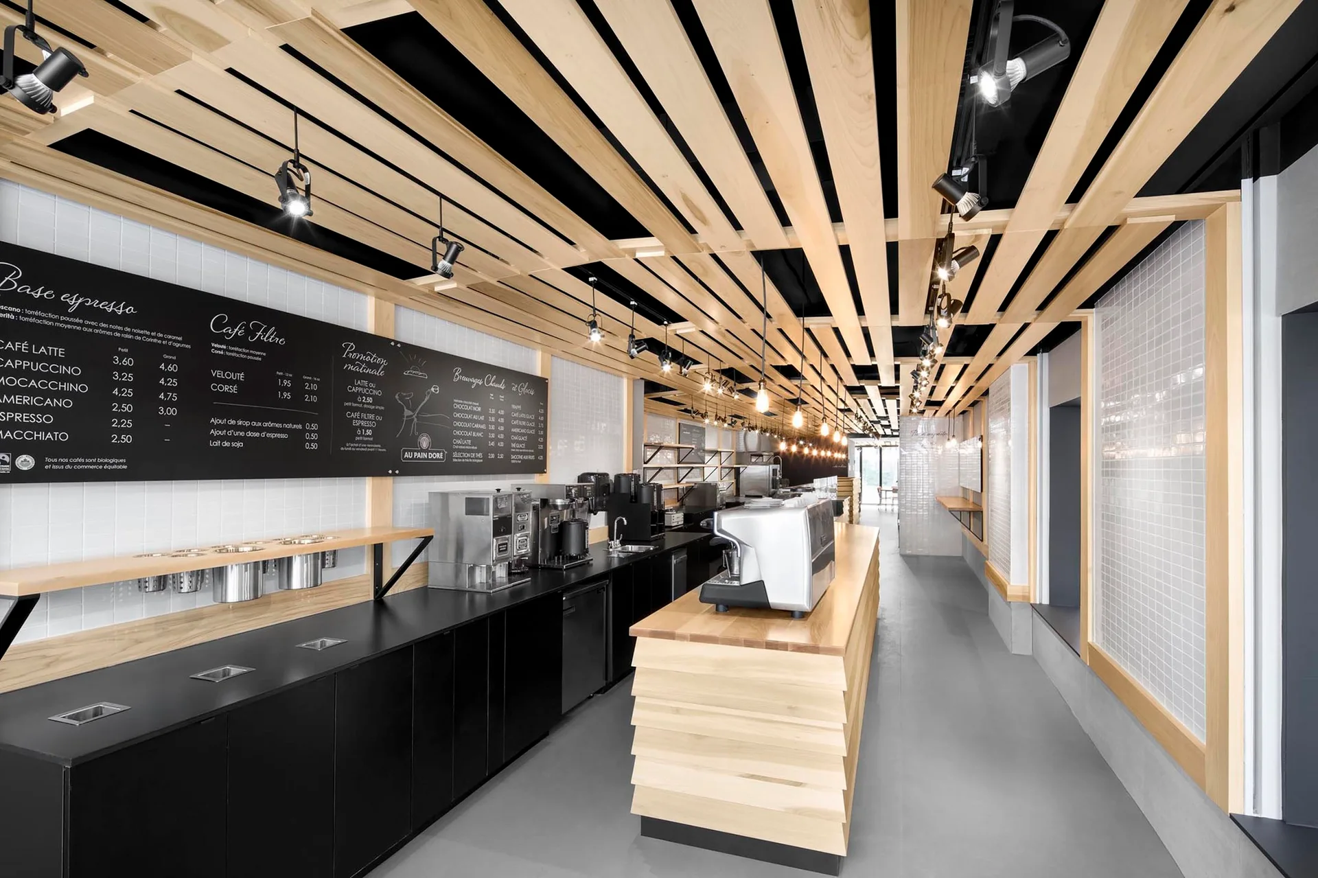 A long commercial cafe interior featuring a light wood service counter, black lower cabinets, white tiled walls, and a linear wood-slat ceiling.